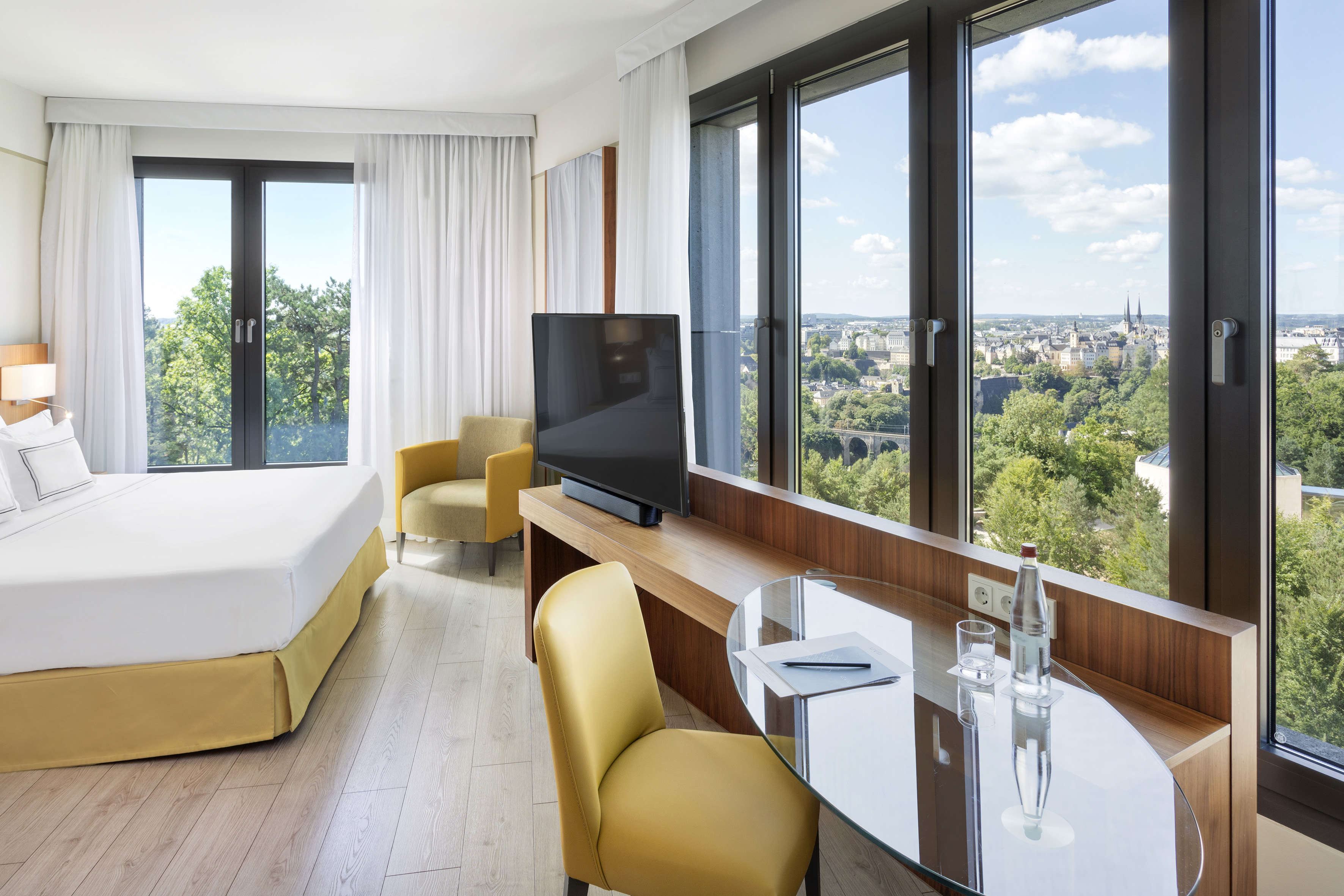 Modern hotel room with panoramic Luxembourg city view, large windows, bed, and desk.