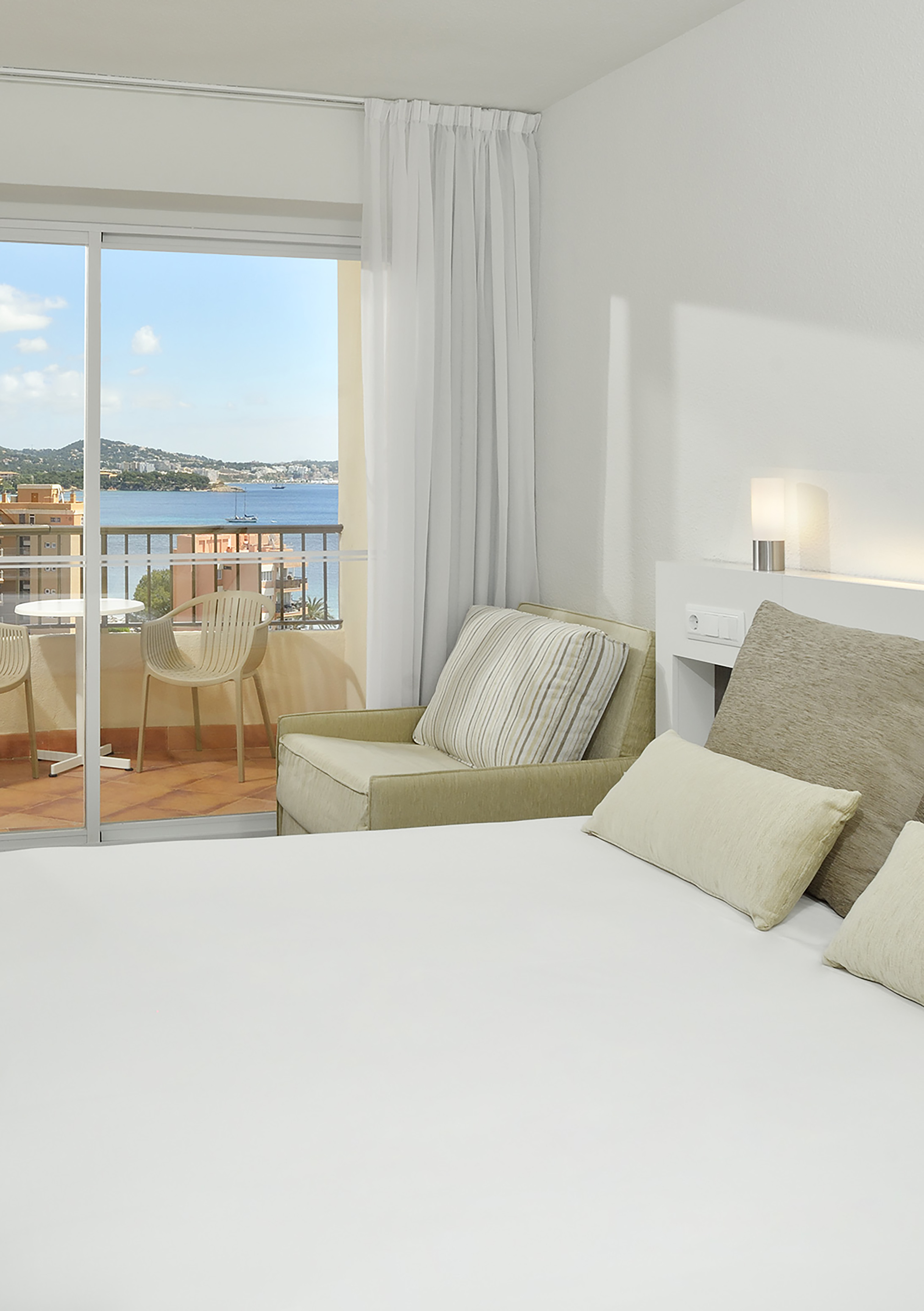 Modern hotel room with balcony, white bed, armchair, and coastal sea view.