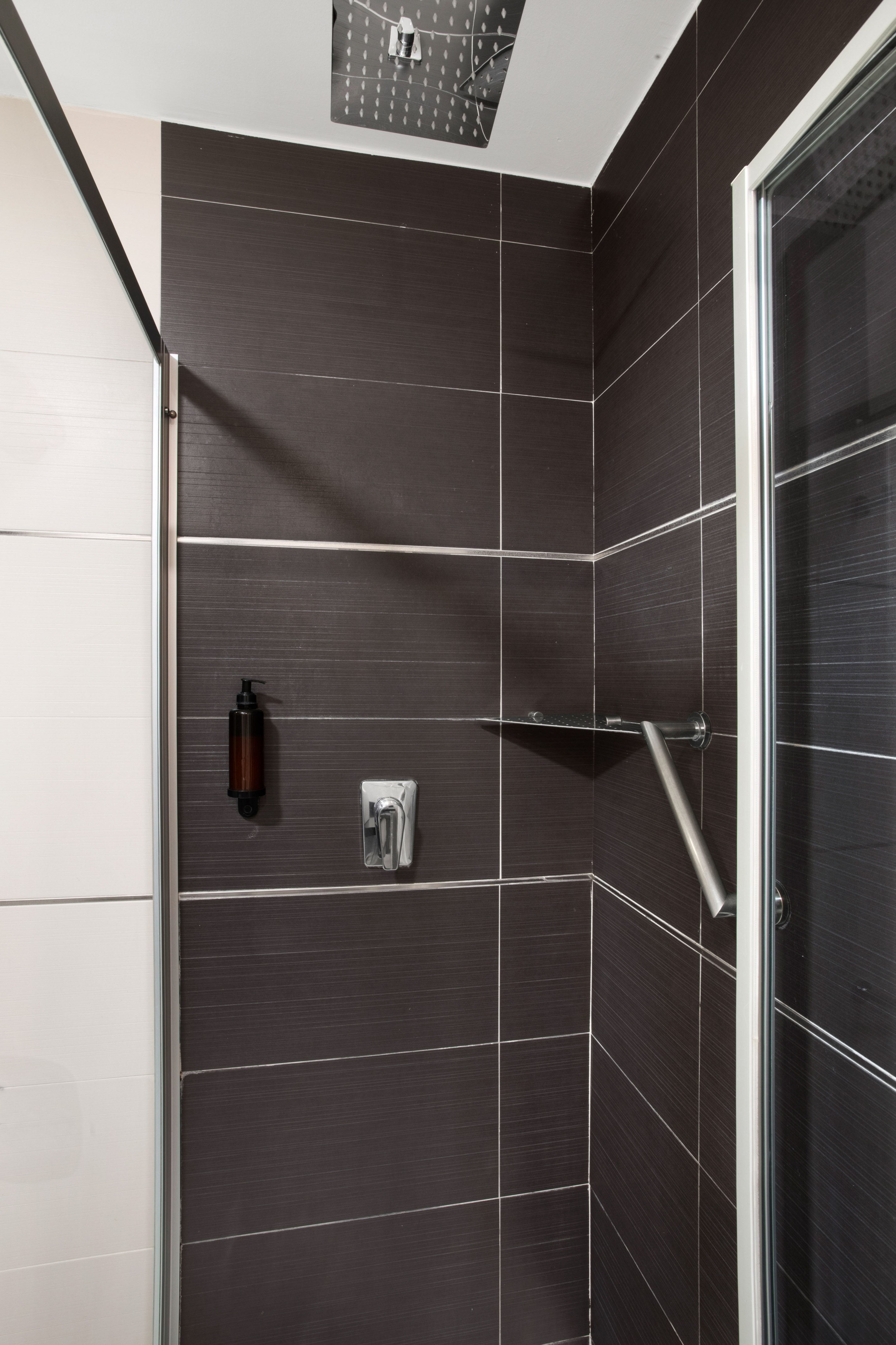 Modern dark tiled shower with rain head, soap dispenser, and safety grab bar.