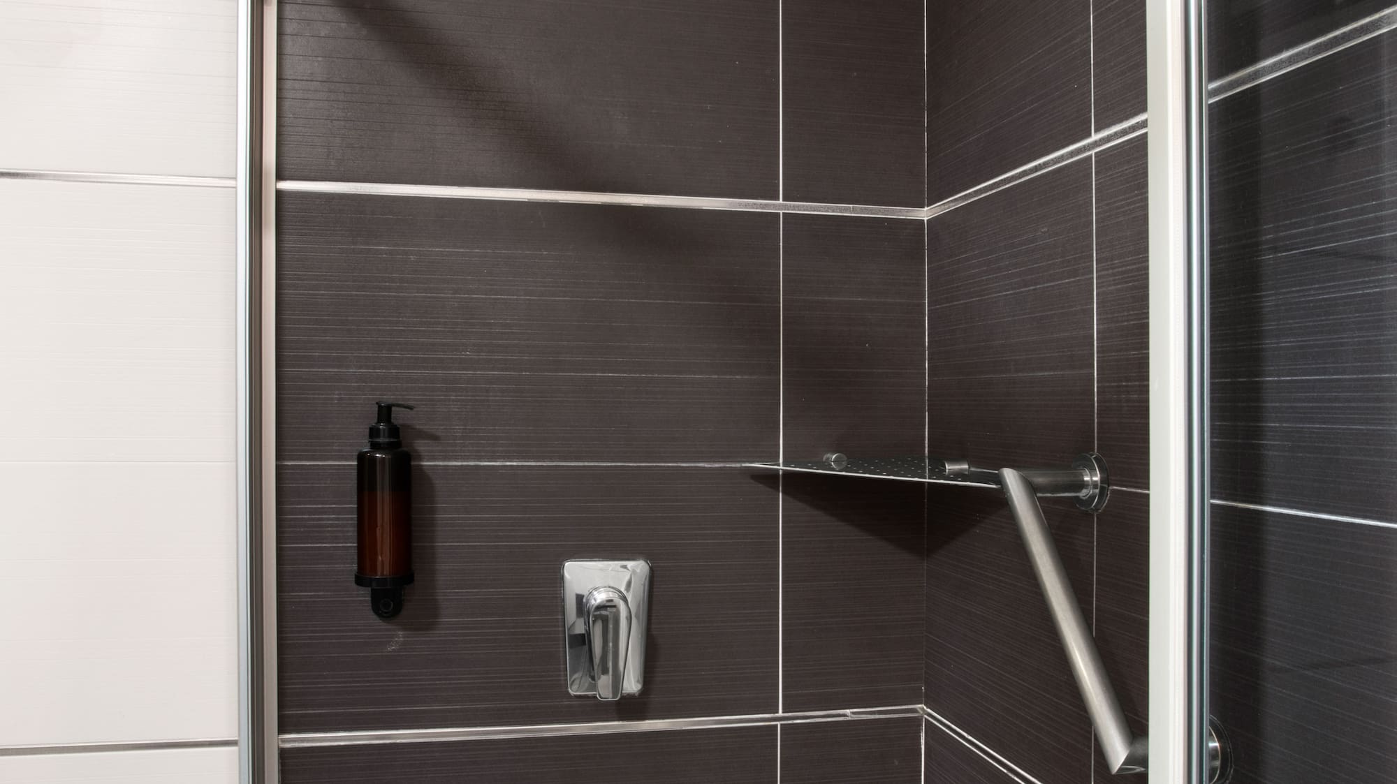 Modern dark tiled shower with rain head, soap dispenser, and safety grab bar.