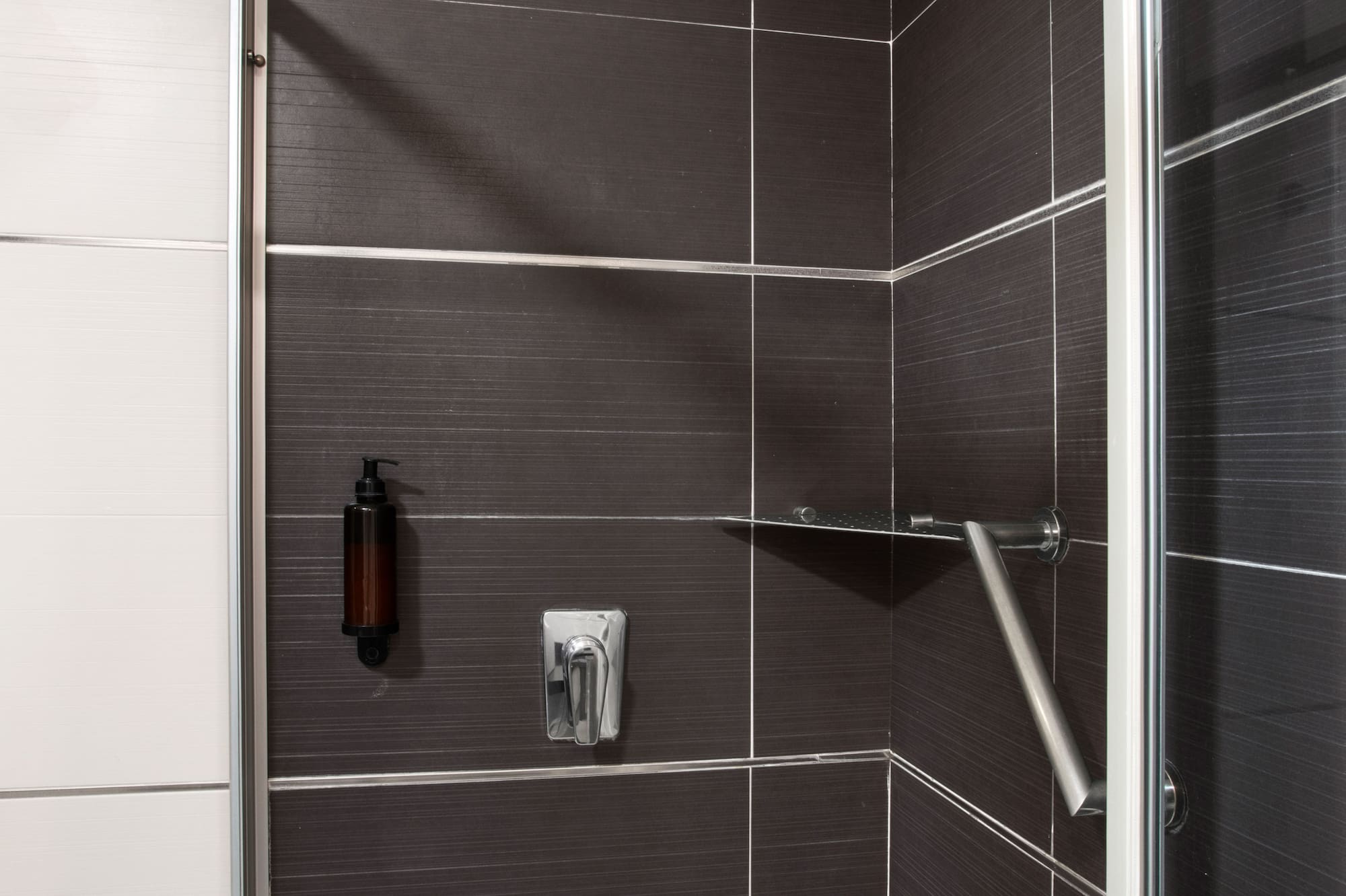 Modern dark tiled shower with rain head, soap dispenser, and safety grab bar.