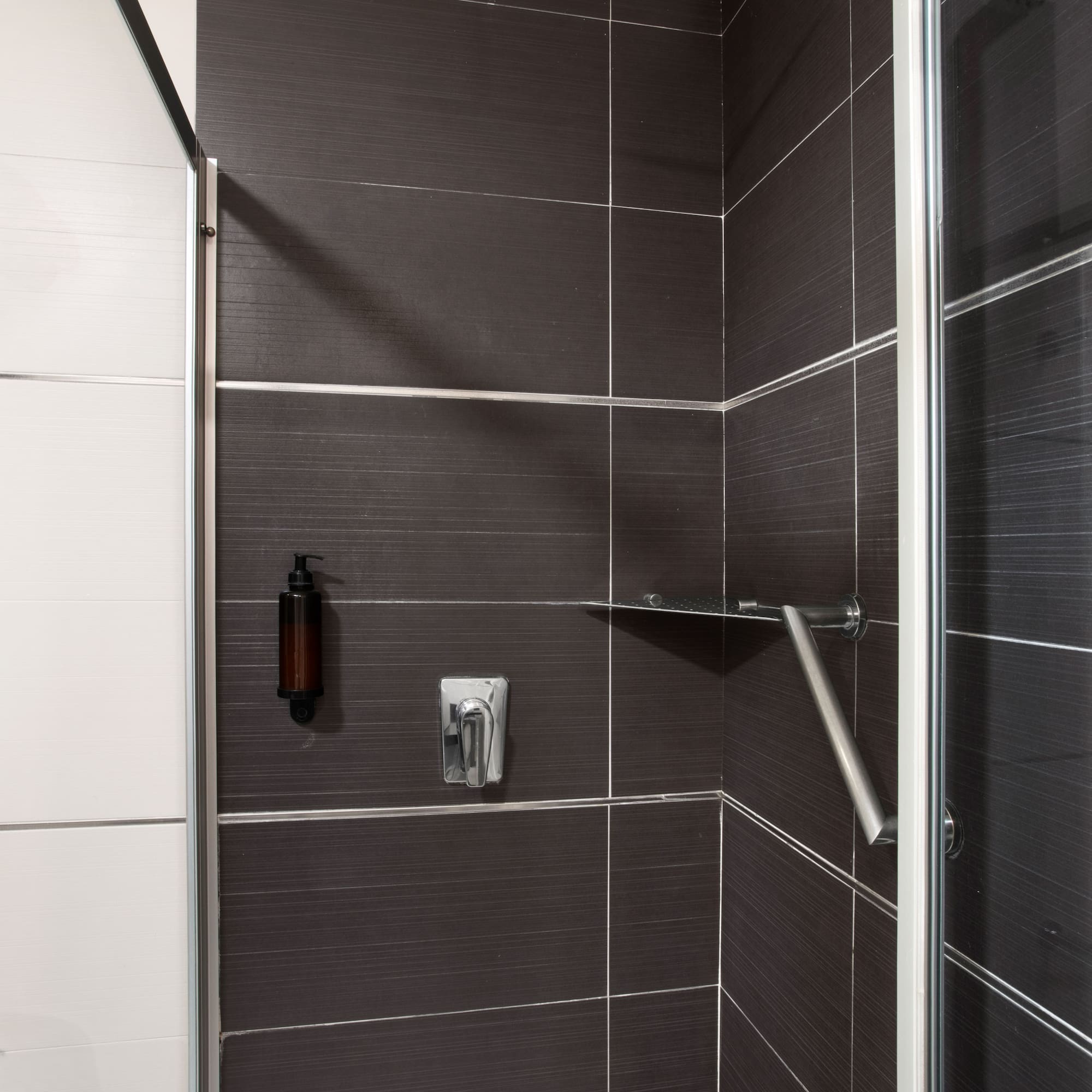 Modern dark tiled shower with rain head, soap dispenser, and safety grab bar.