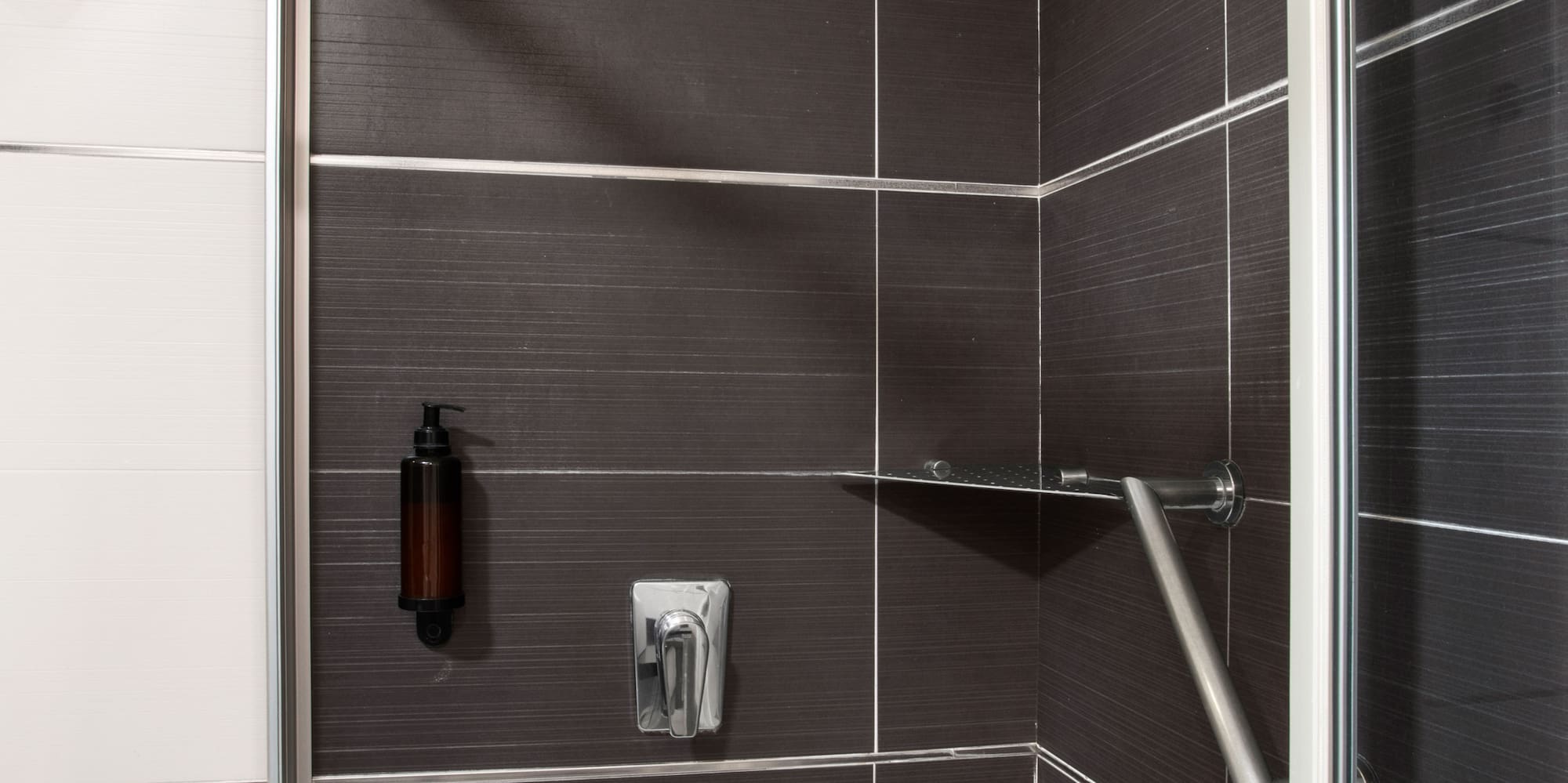 Modern dark tiled shower with rain head, soap dispenser, and safety grab bar.