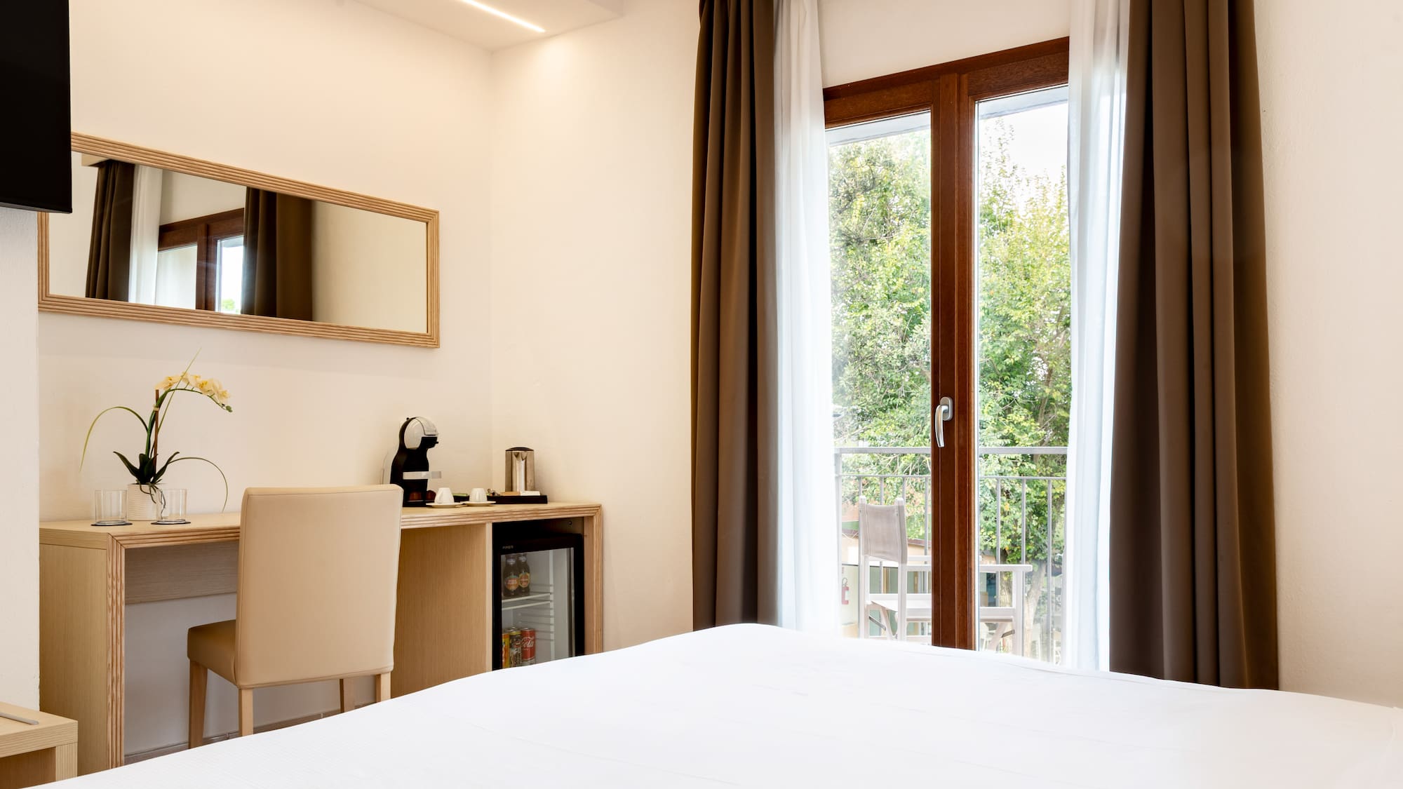 Hotel room with white bed, balcony, desk, Nespresso machine, and mini-bar.