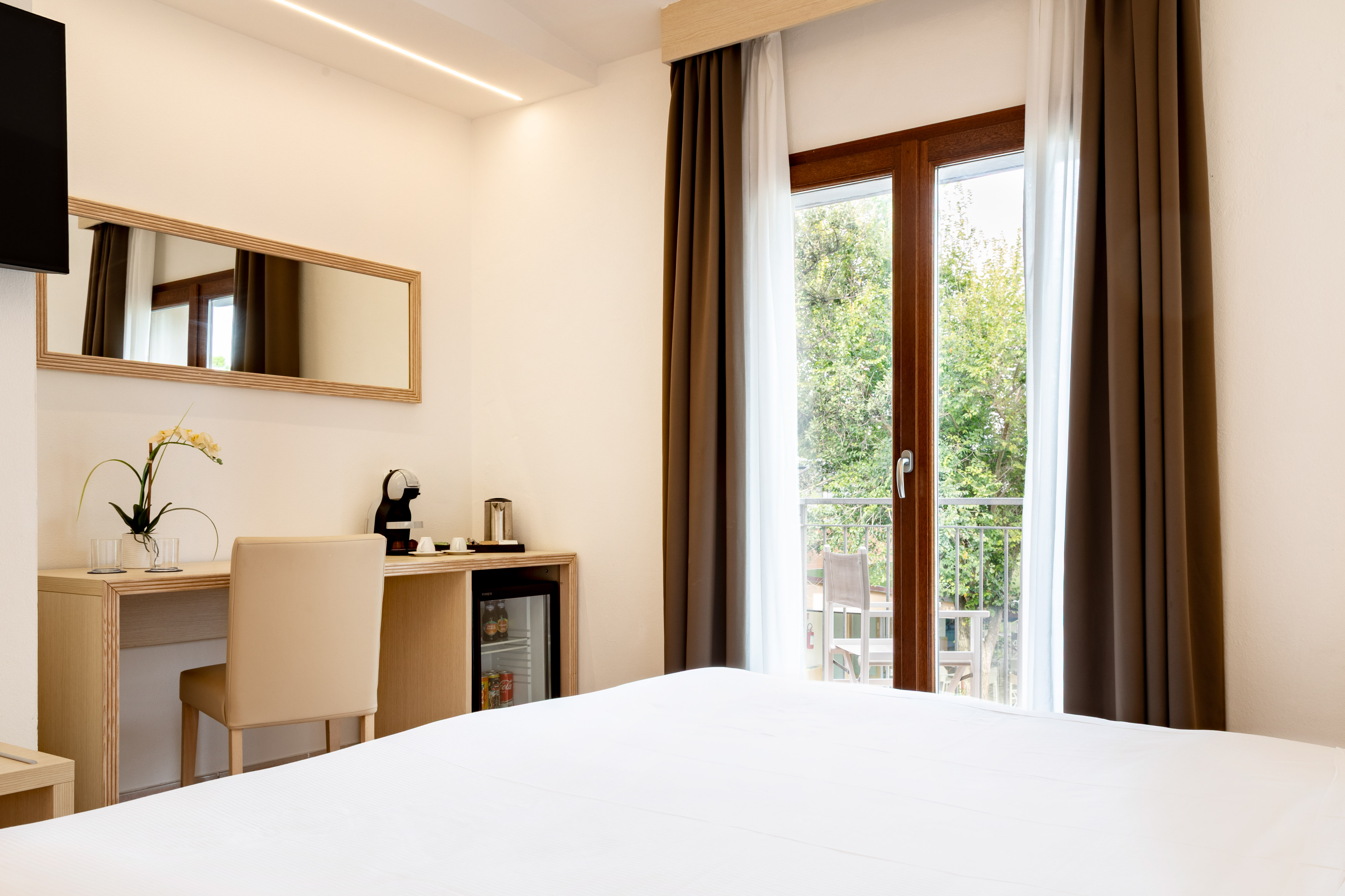 Hotel room with white bed, balcony, desk, Nespresso machine, and mini-bar.