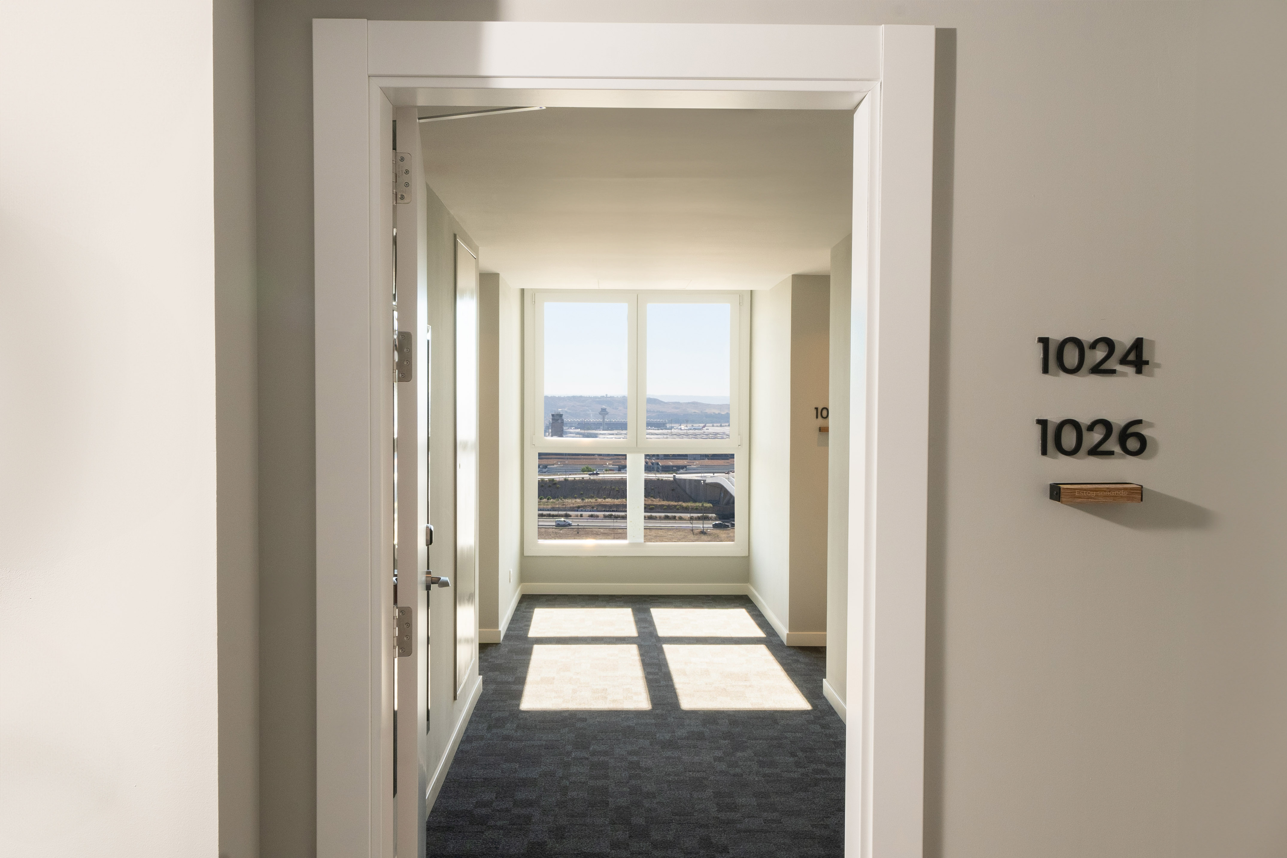 Modern corridor with large window, airport view, sunlit carpet. Room numbers 1024, 1026.