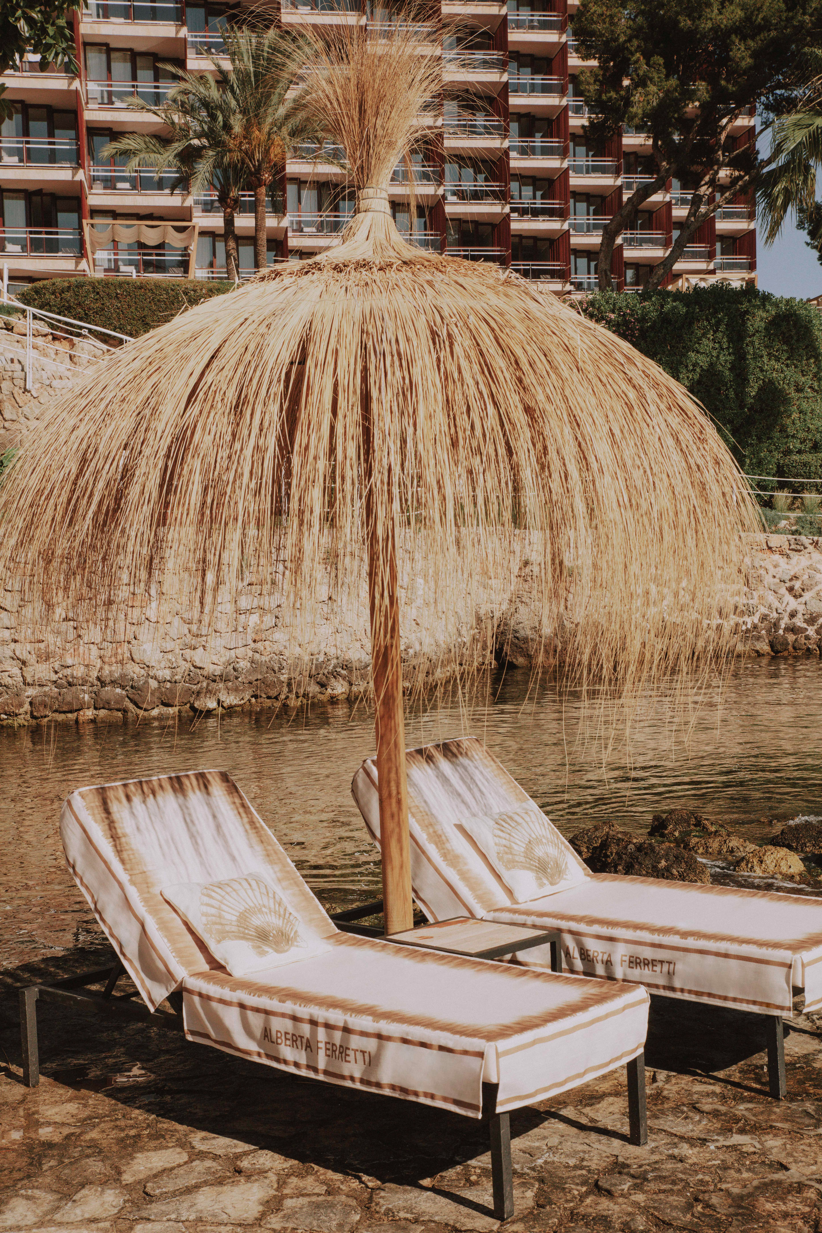 Alberta Ferretti lounge chairs under straw umbrella by hotel on calm bay.