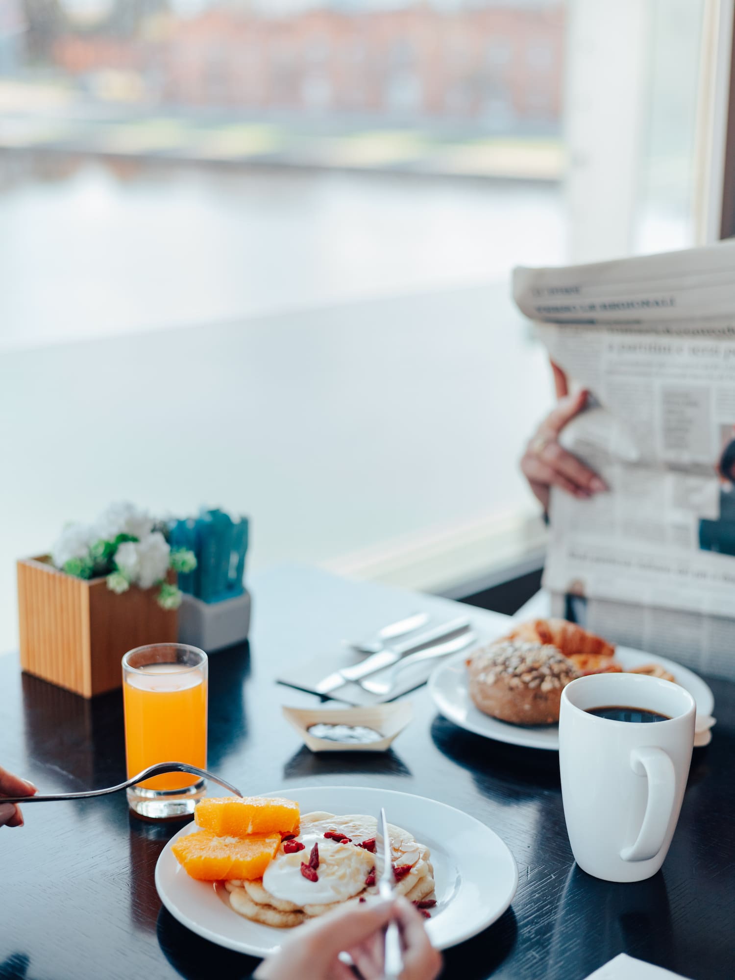 Hotel breakfast with pancakes, orange juice, coffee, and newspaper by a window.