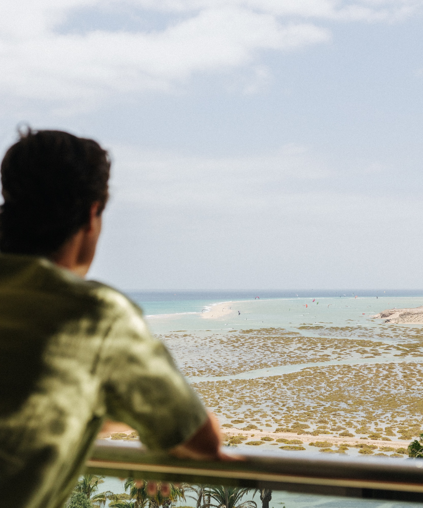 Man viewing coastal lagoon, ocean, and windsurfers from balcony.