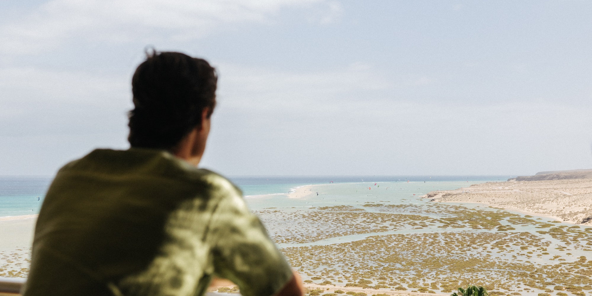 Man on balcony overlooks coastal lagoon, ocean, and windsurfers.