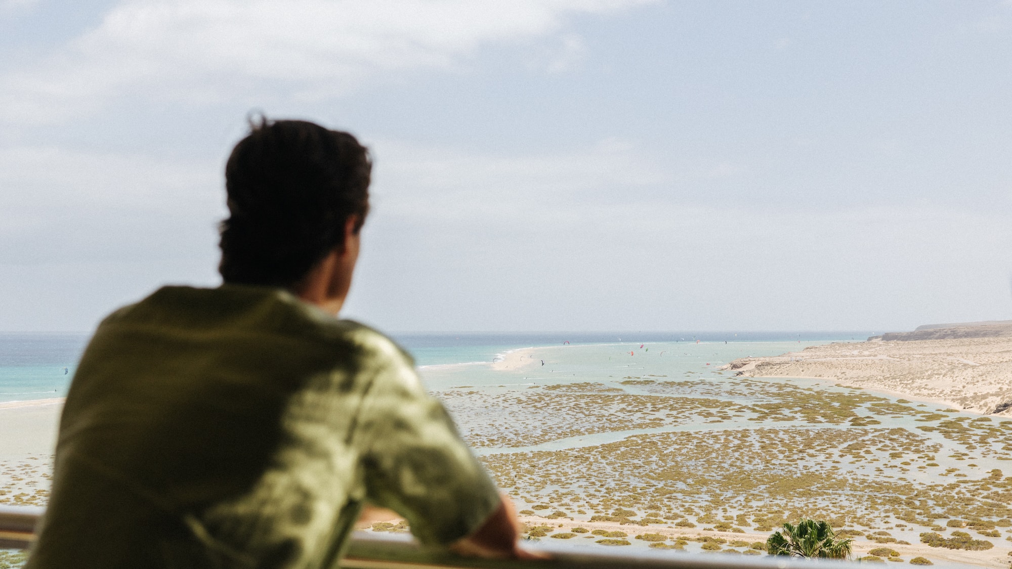 Man on balcony overlooks coastal lagoon, ocean, and windsurfers.