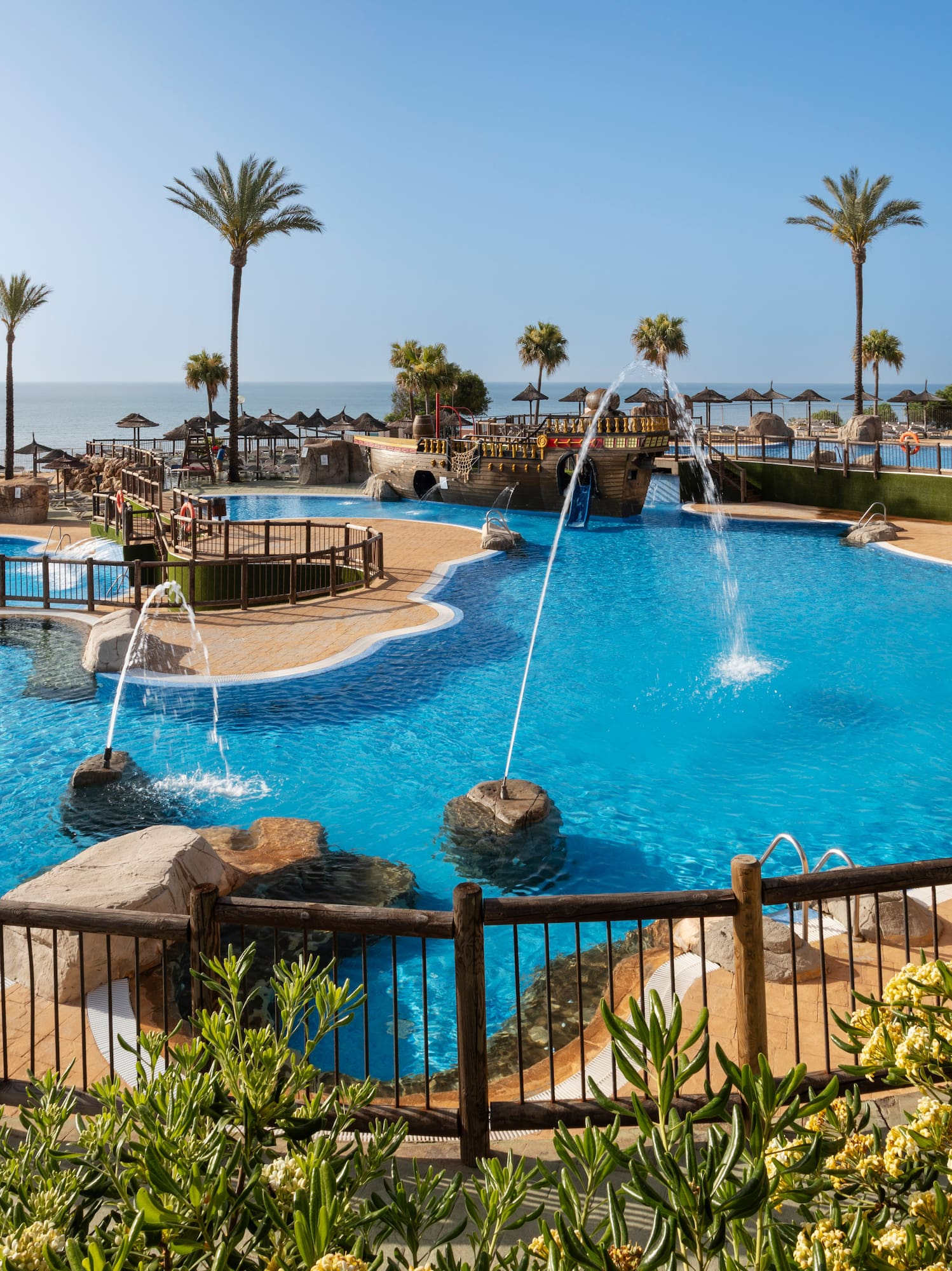 Resort swimming pool with fountains, palm trees, beach, and ocean view.