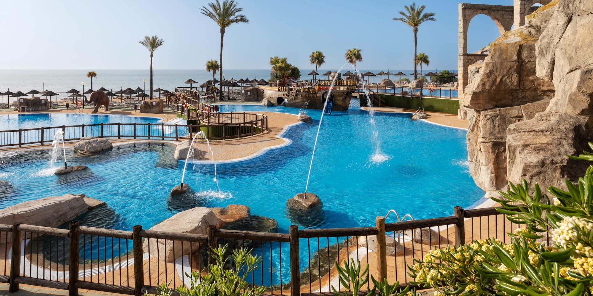 Resort water park pool with fountains, palm trees, beach, and ocean view.