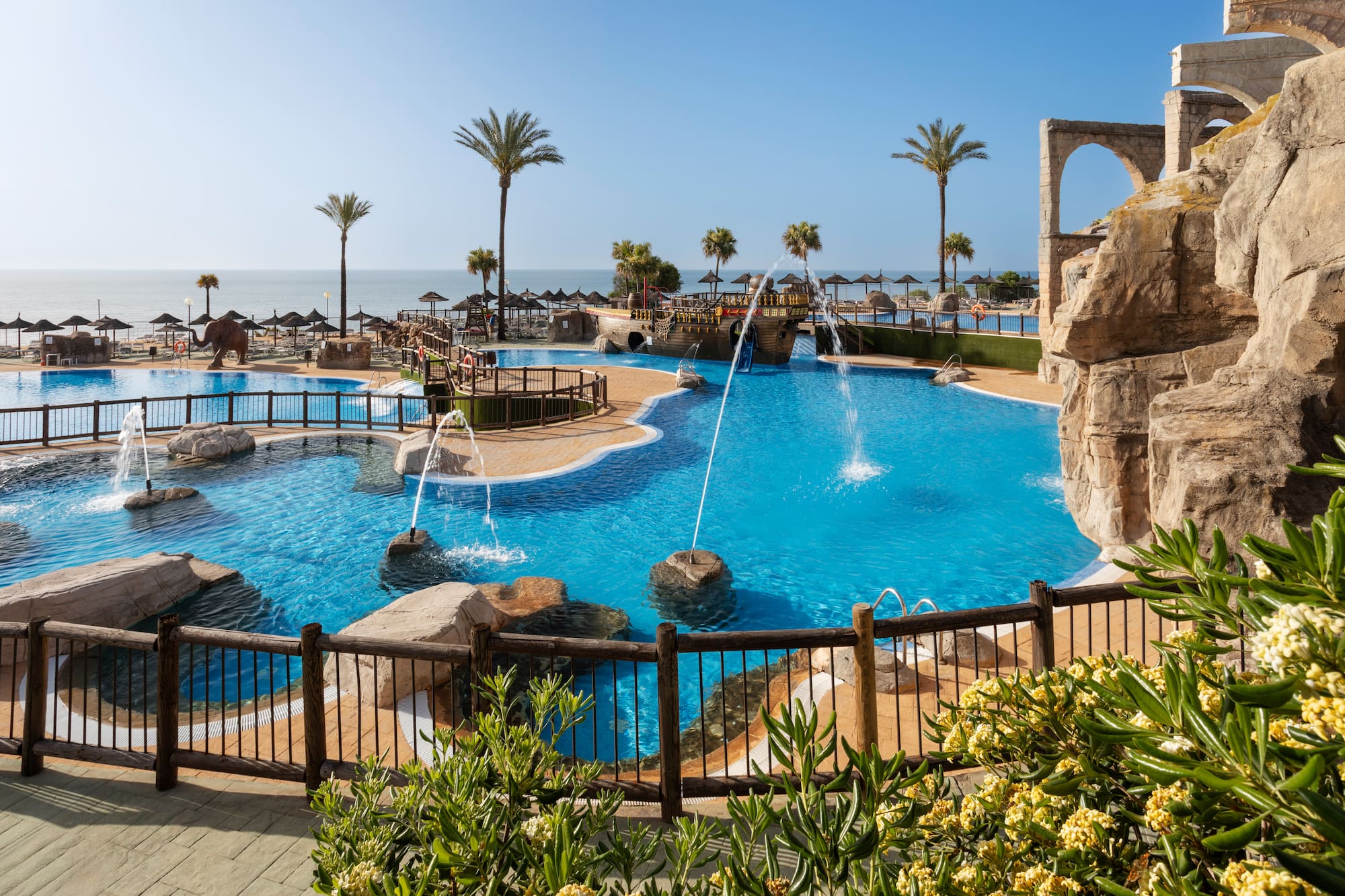 Resort water park pool with fountains, palm trees, beach, and ocean view.