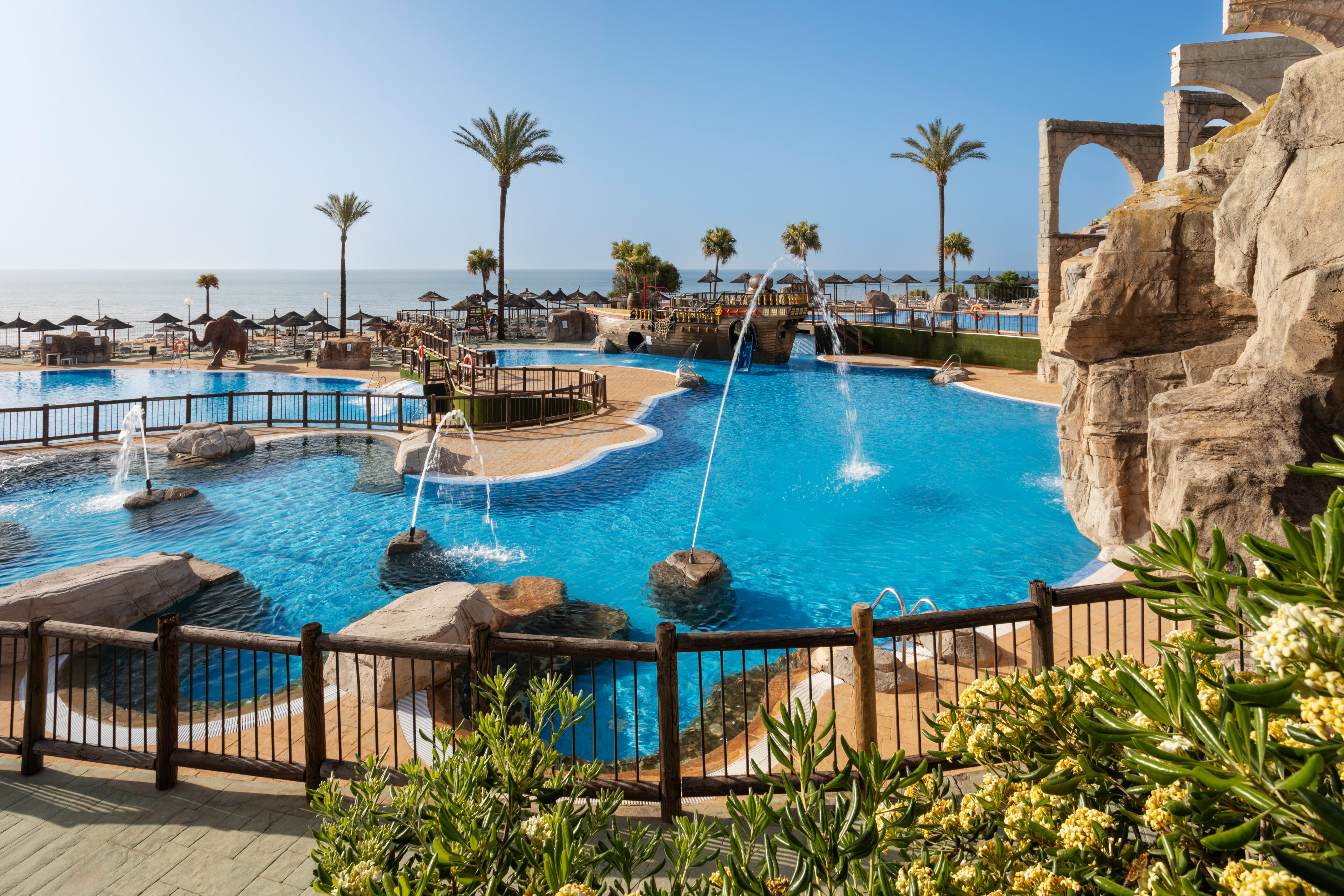Resort swimming pool with fountains, palm trees, beach, and ocean view.