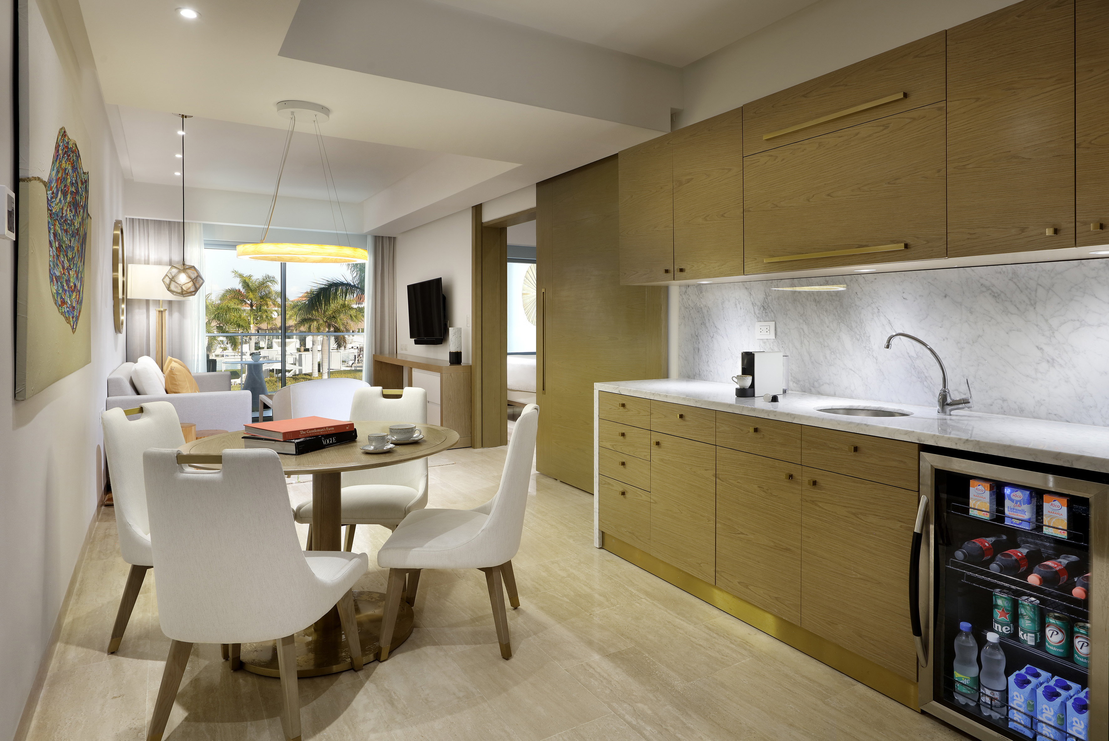 Modern hotel suite with kitchenette, dining, living area, and tropical view.