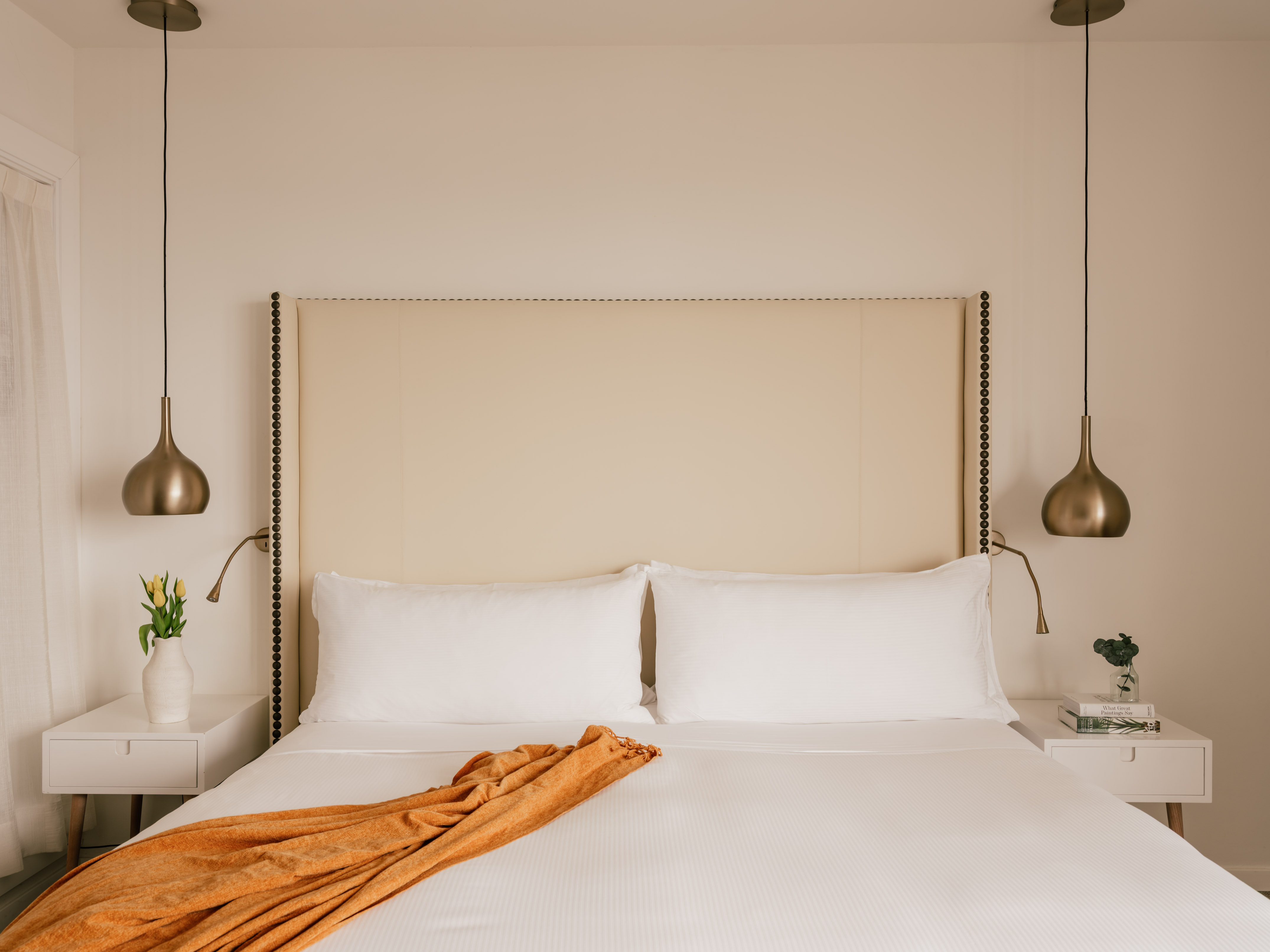Hotel bedroom with upholstered headboard, white bed, orange throw, brass lights.