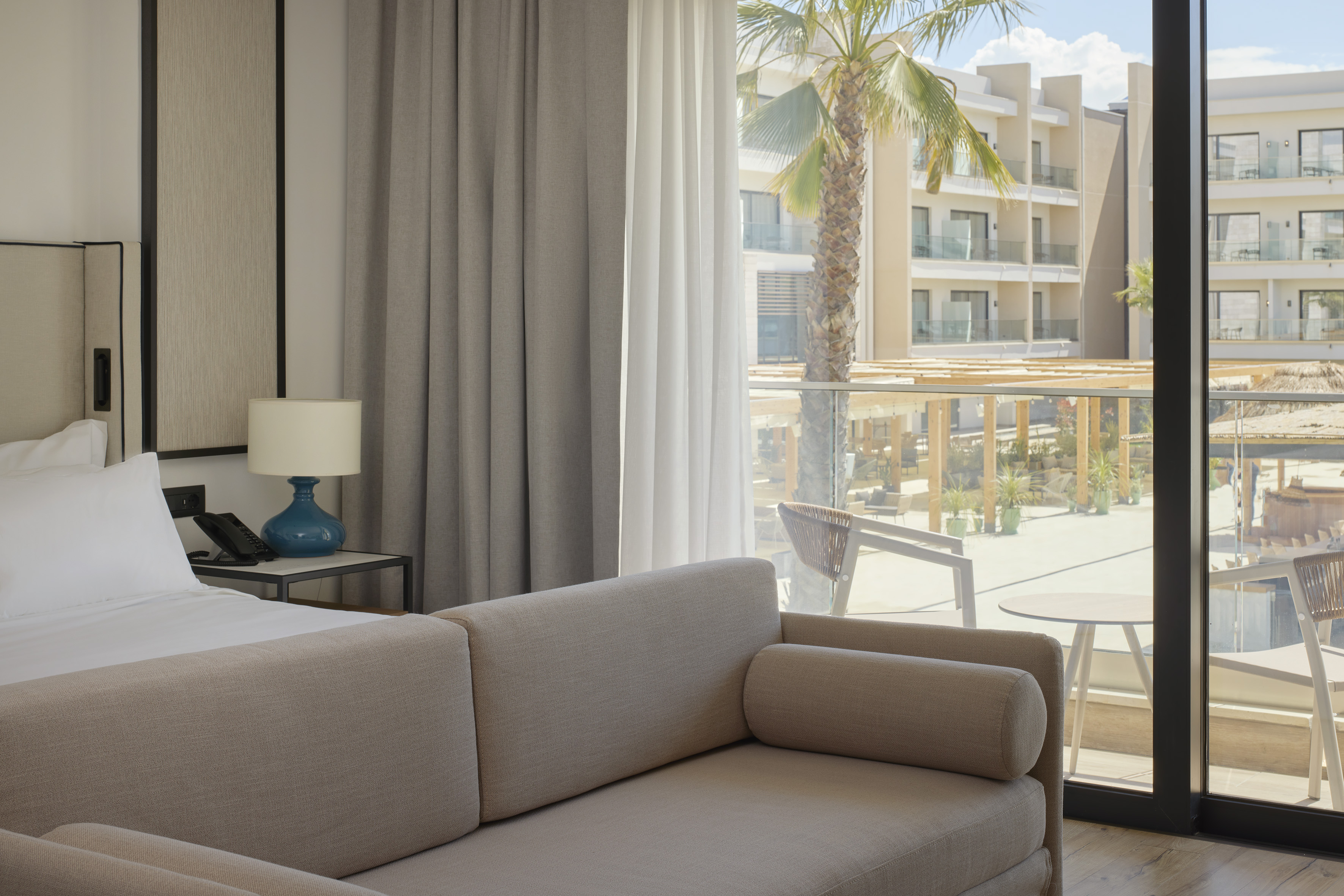 Bright hotel room with sofa, bed, and balcony view of resort and palm trees.
