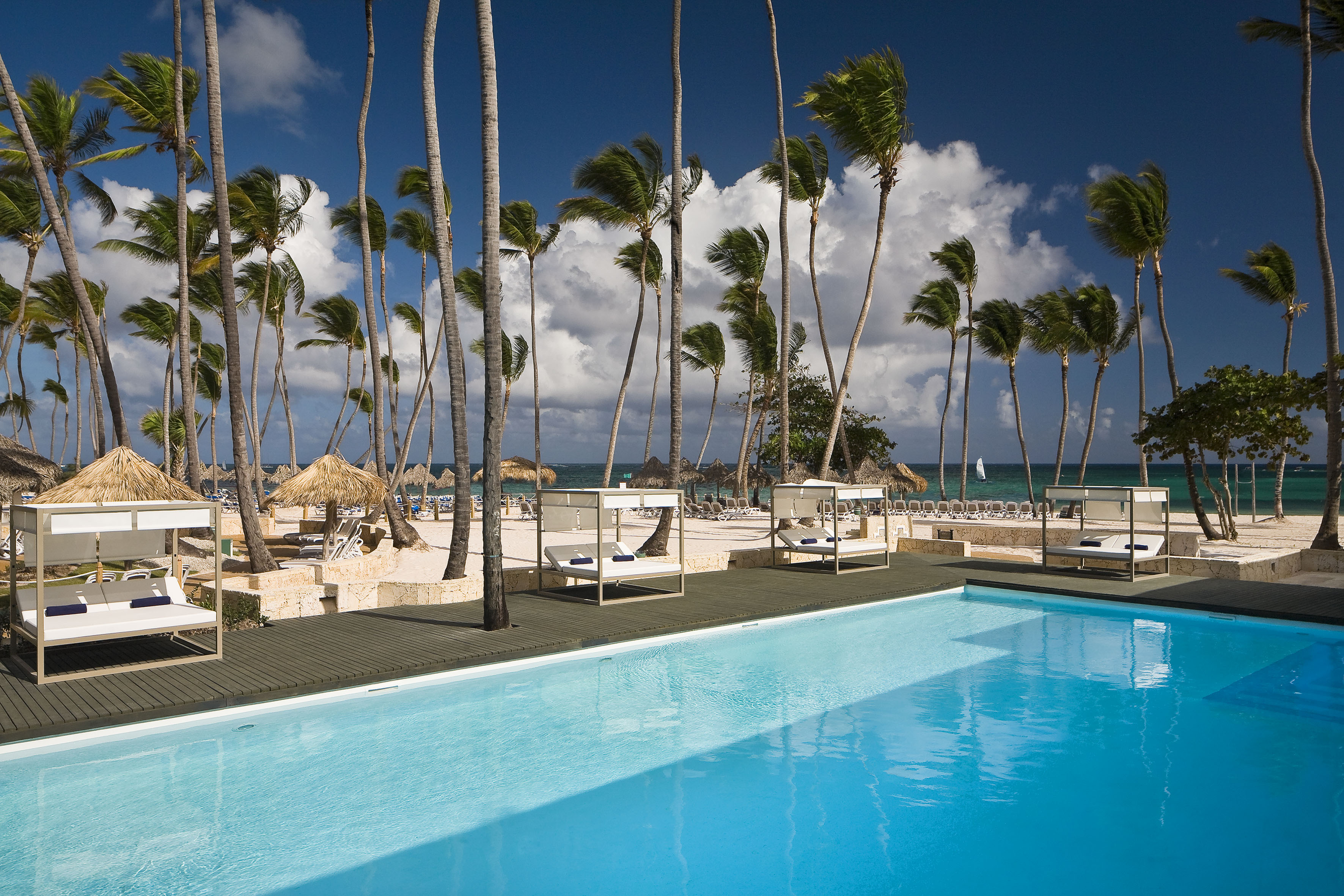 Tropical resort with blue pool, palm trees, beach cabanas, and ocean.