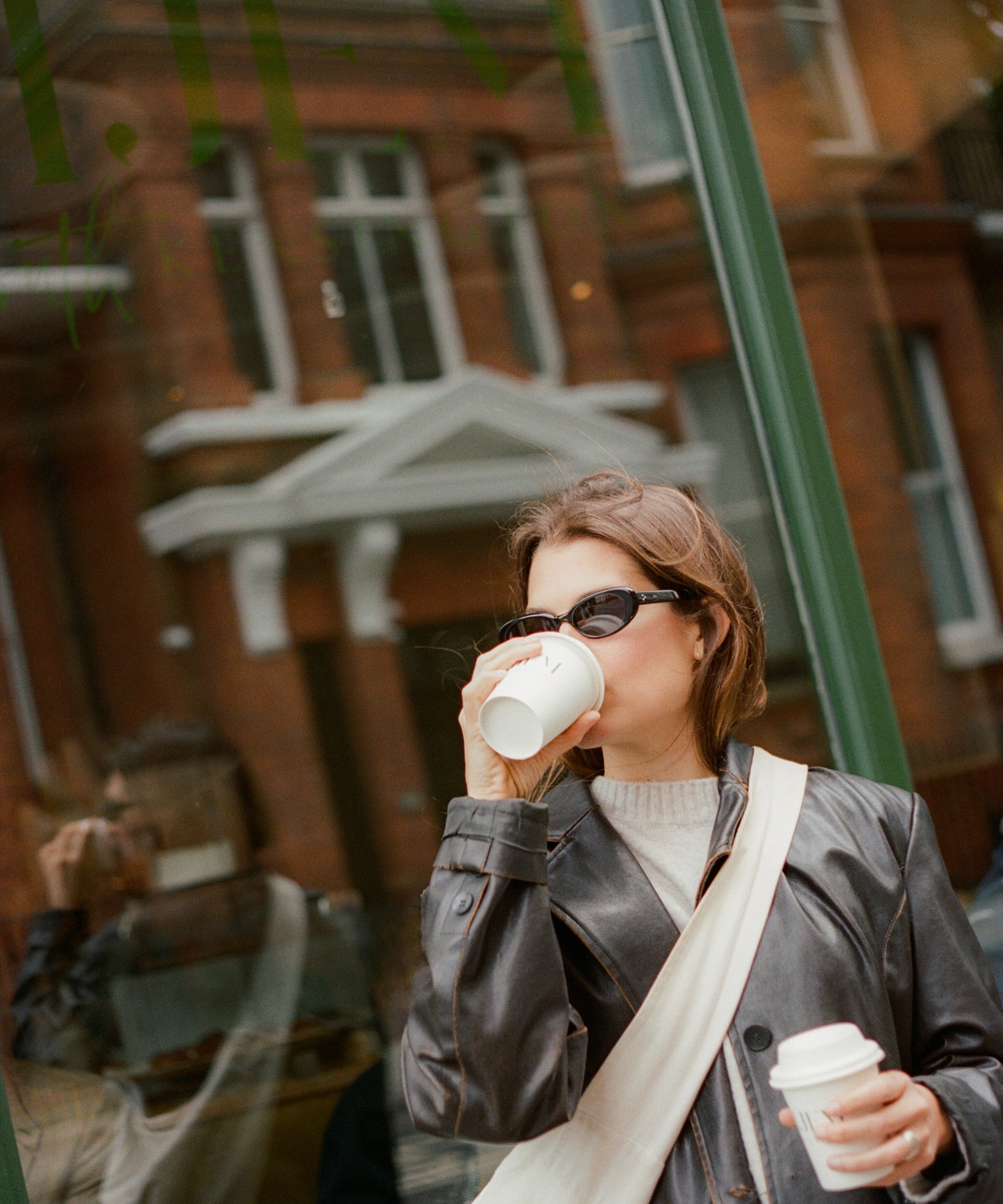 Mujer con chaqueta de cuero, gafas de sol y café frente a ventana urbana.