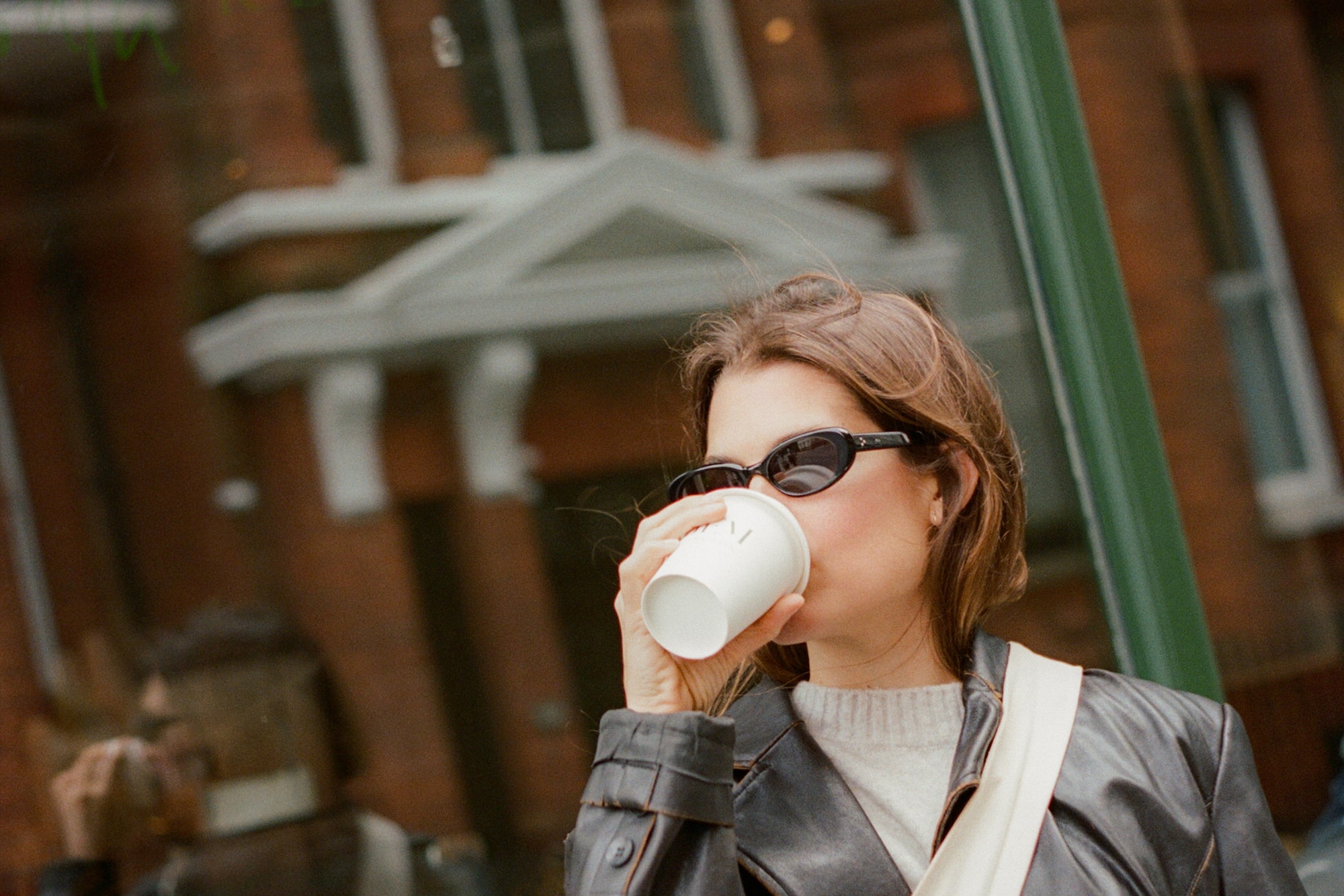 Mujer con chaqueta de cuero, gafas de sol y café frente a ventana urbana.