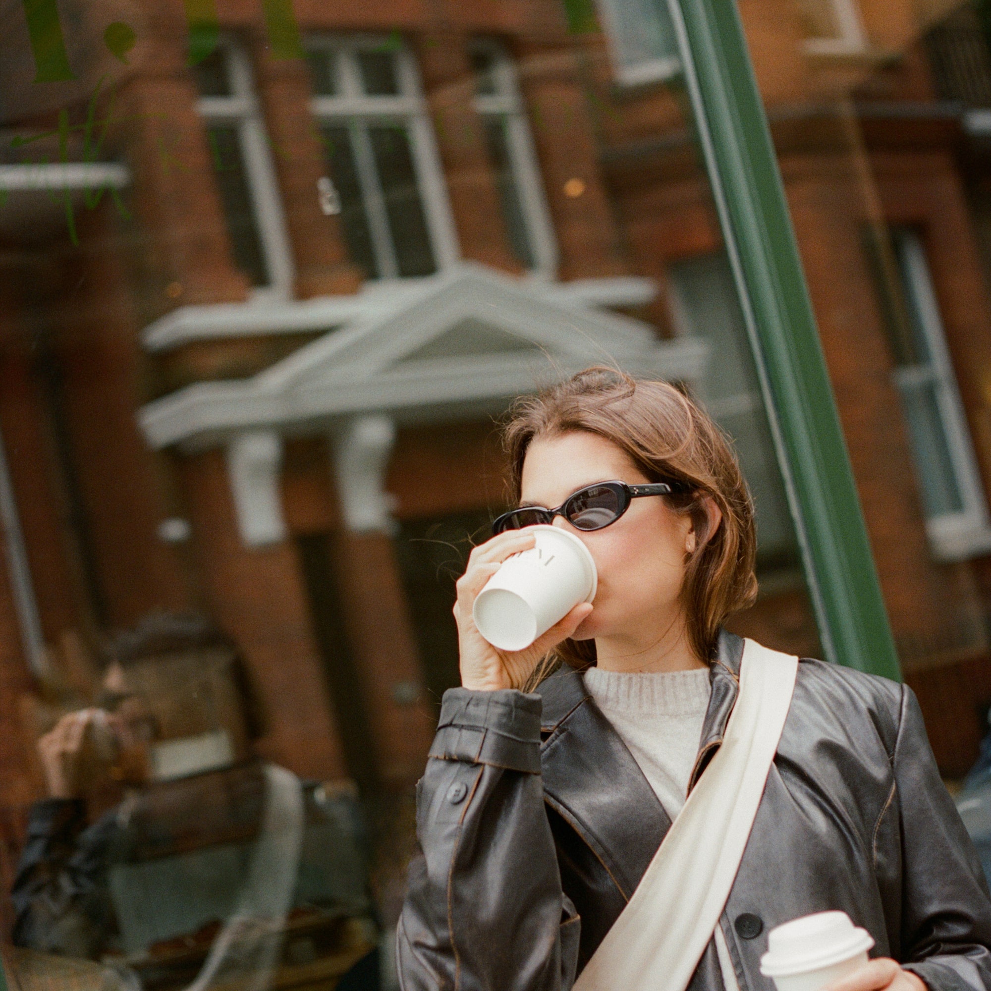 Mujer con chaqueta de cuero, gafas de sol y café frente a ventana urbana.