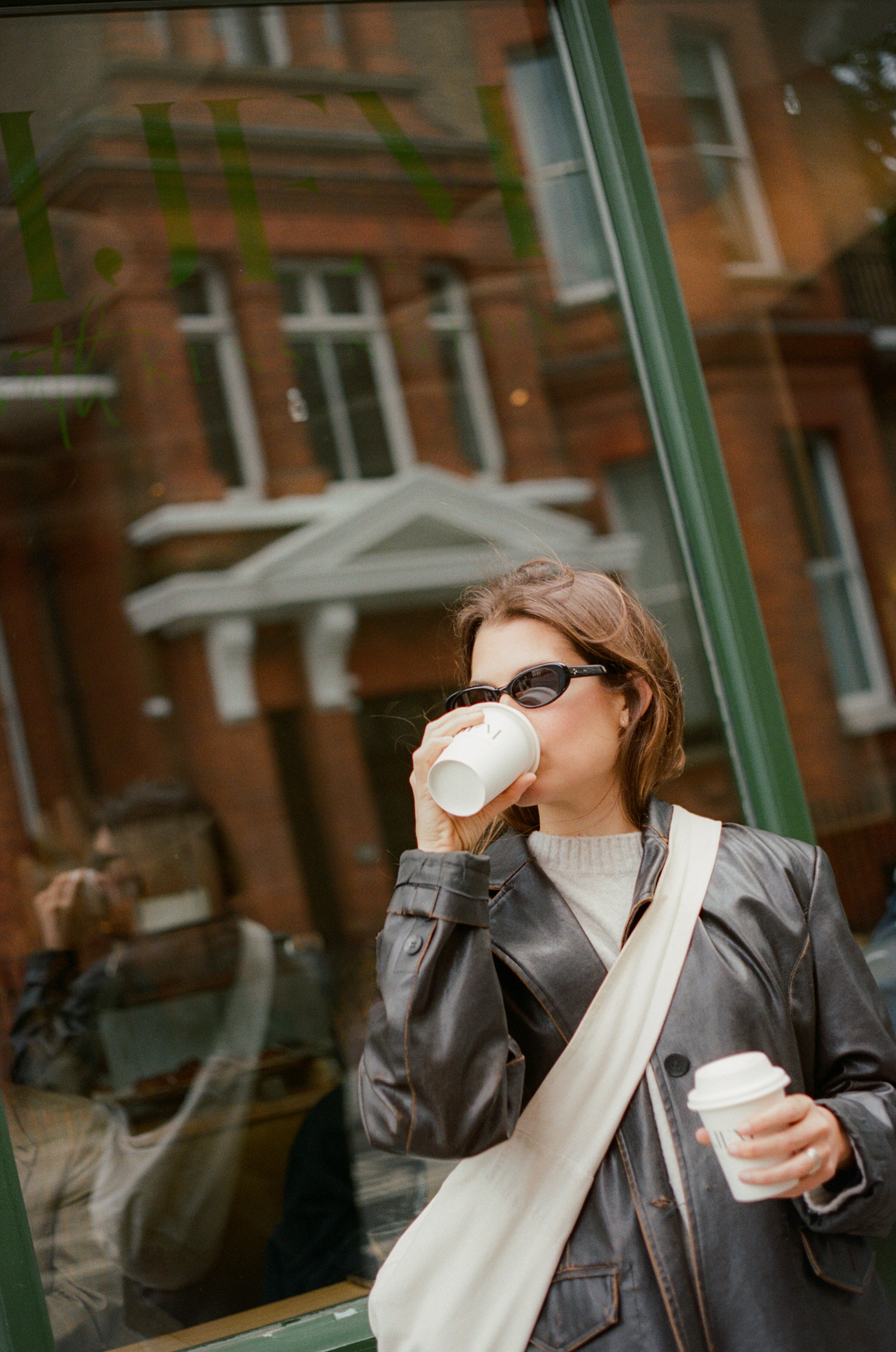 Mujer con chaqueta de cuero, gafas de sol y café frente a ventana urbana.