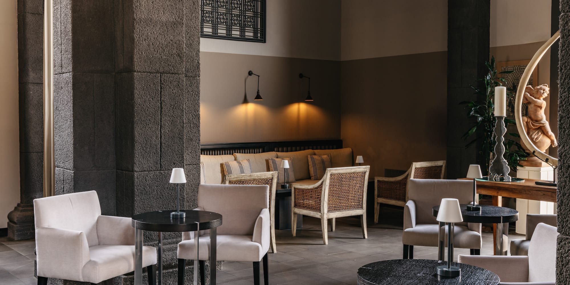 Cozy hotel lounge with stone columns, armchairs, dark tables, and warm lighting.