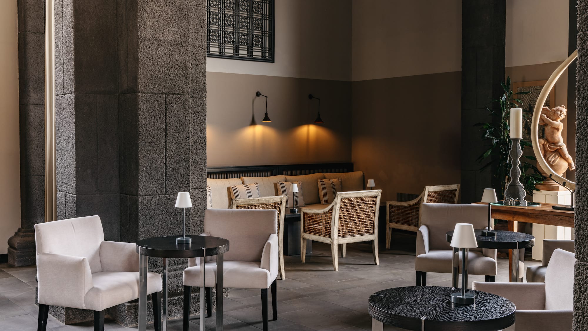 Cozy hotel lounge with stone columns, armchairs, dark tables, and warm lighting.