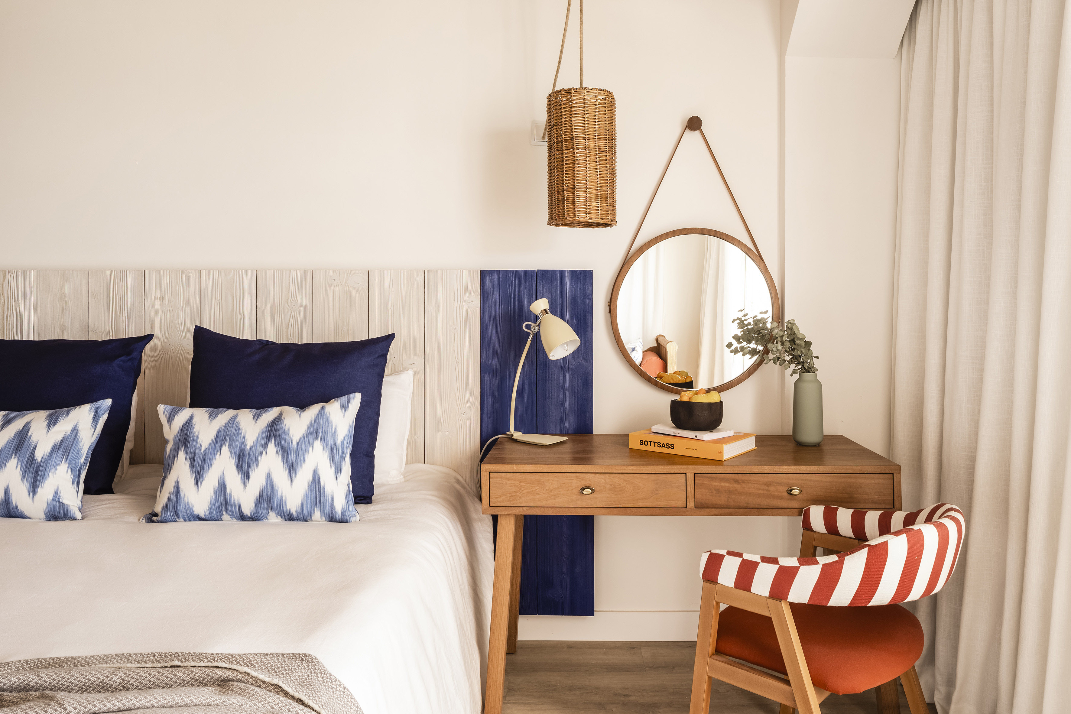 Modern bedroom: white bed, blue pillows, wood desk, round mirror, striped chair.