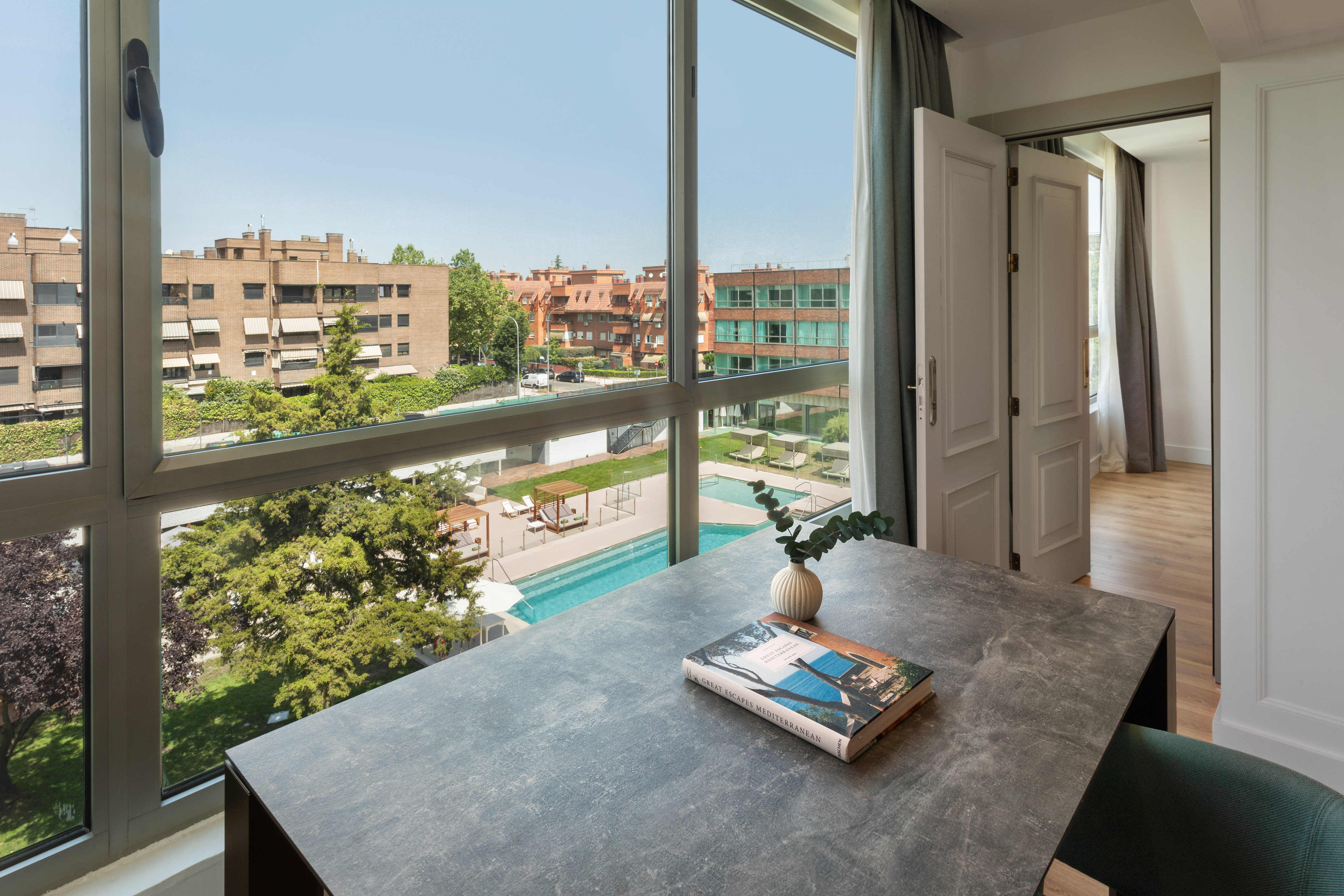 Apartment view: modern desk, large window, sunny pool area, residential buildings.