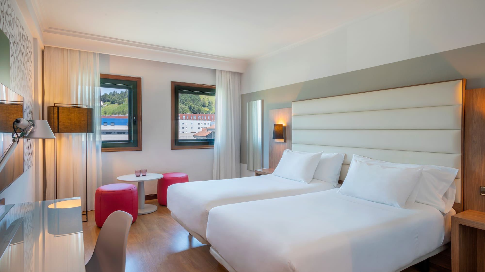 Modern hotel room with twin beds, desk, red stools, and window view.