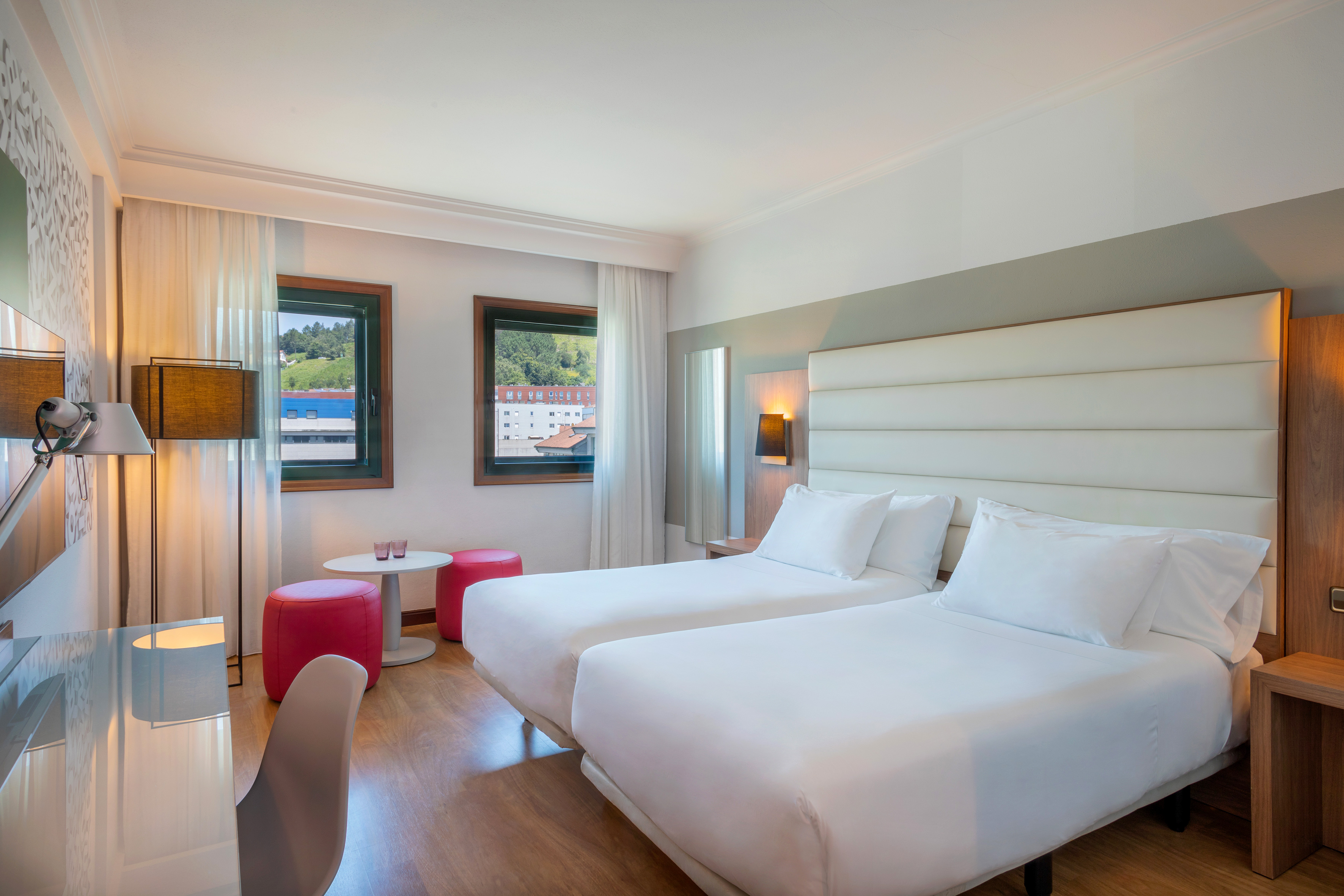 Modern hotel room with twin beds, desk, red stools, and window view.