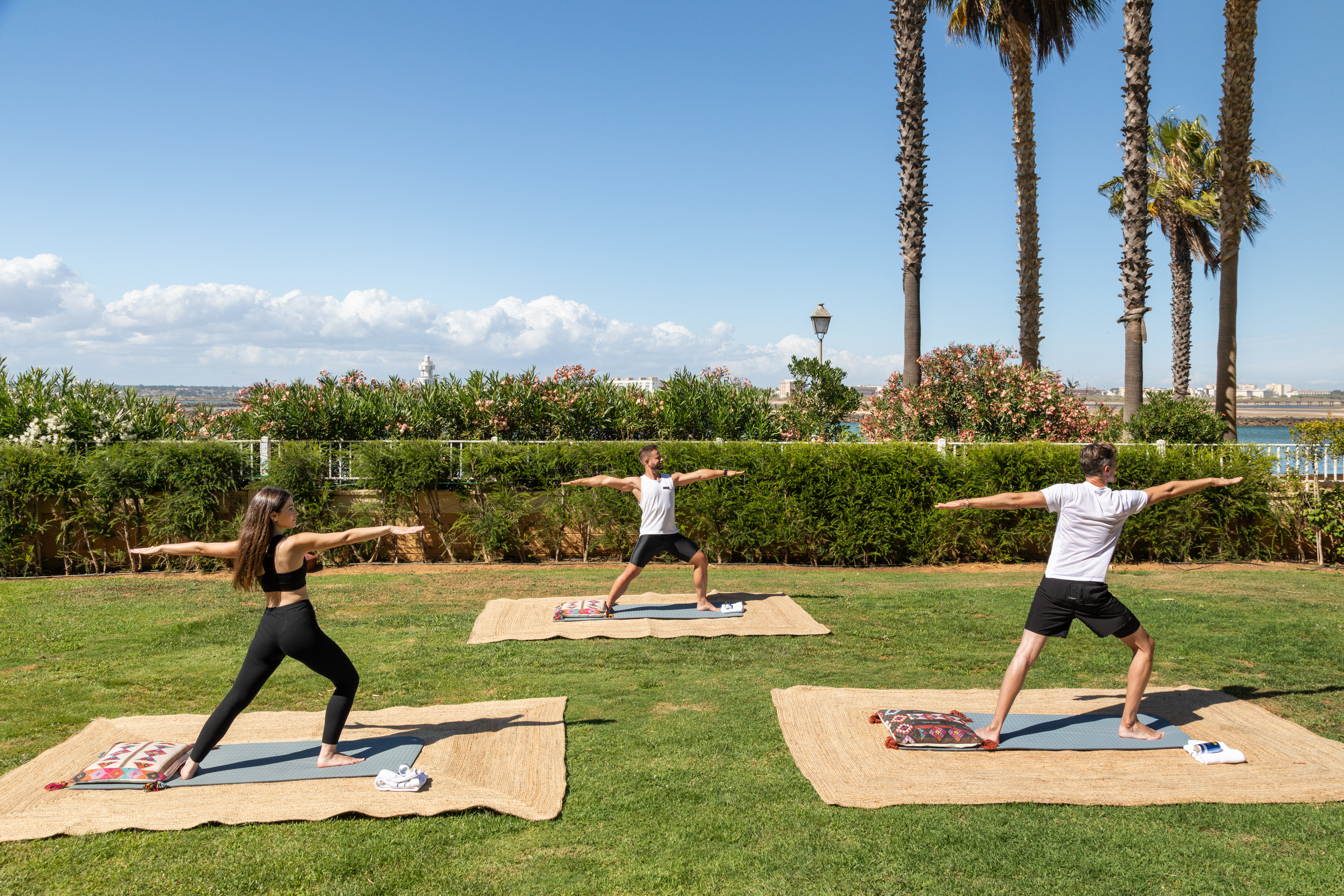 Sunny outdoor yoga with three people on mats in a tropical garden by the sea.