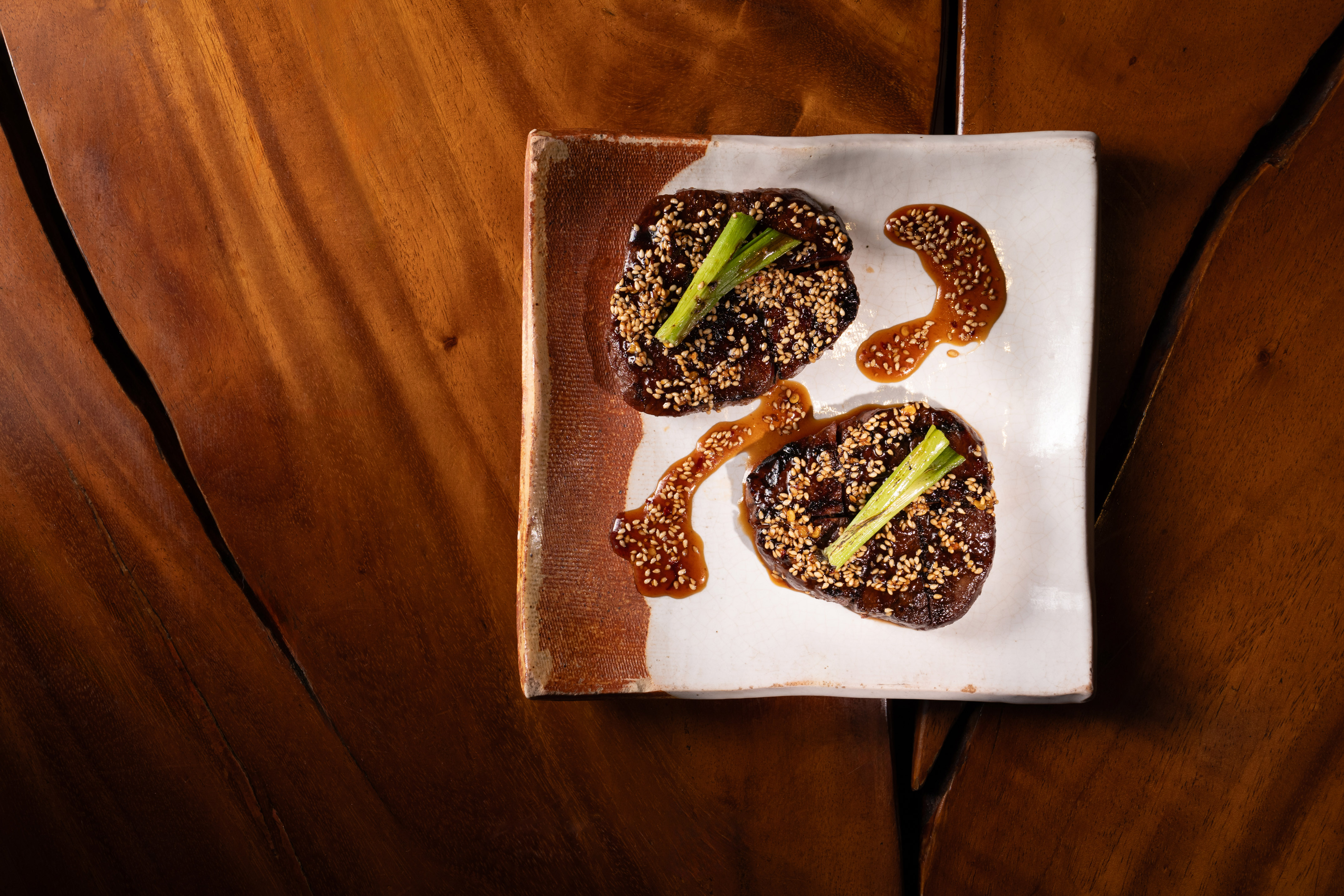 Glazed beef medallions with sesame seeds, green onion, and sauce on rustic plate.