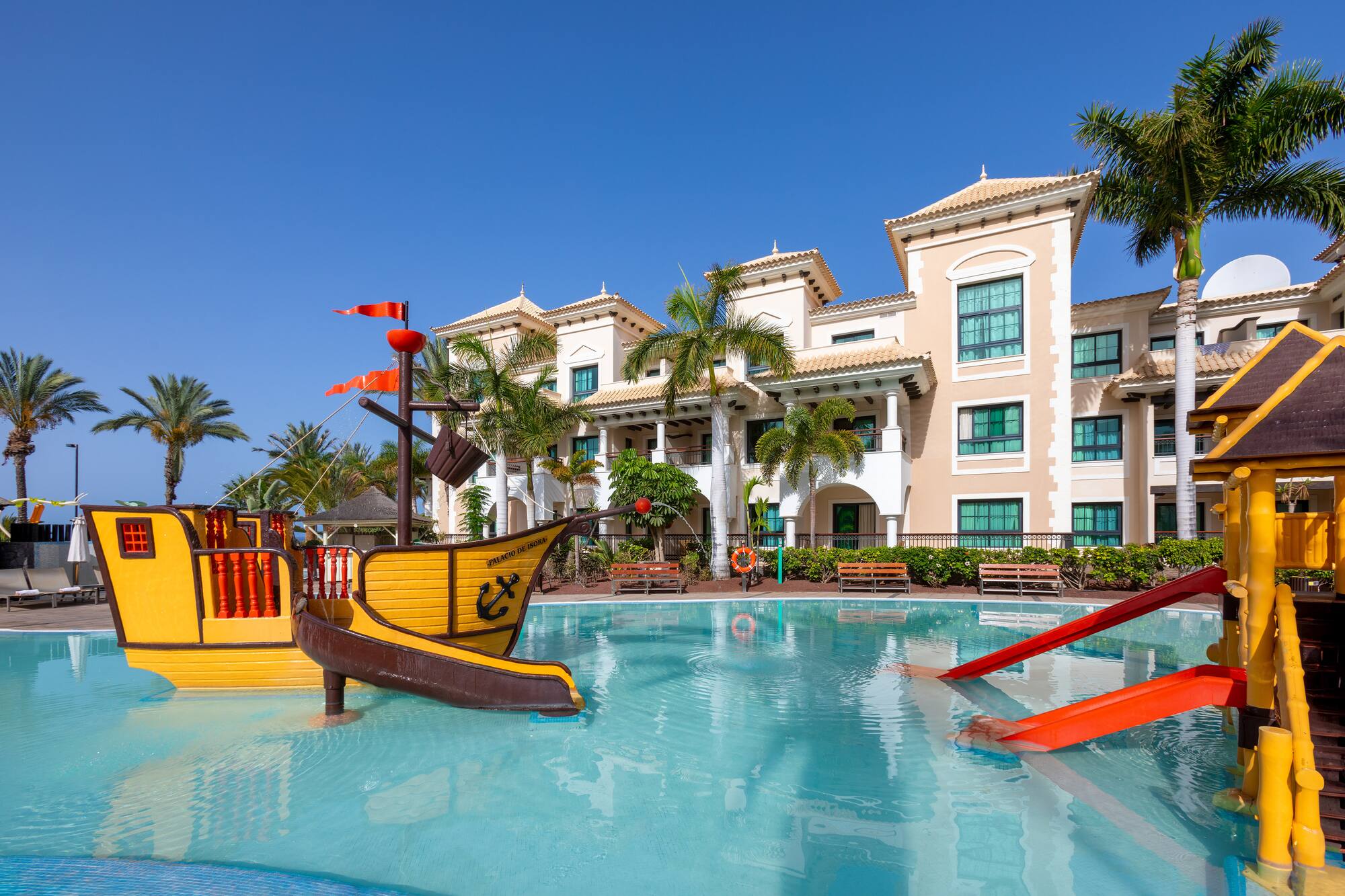 Resort kids' pool with pirate ship, water slides, tropical hotel, and palm trees.
