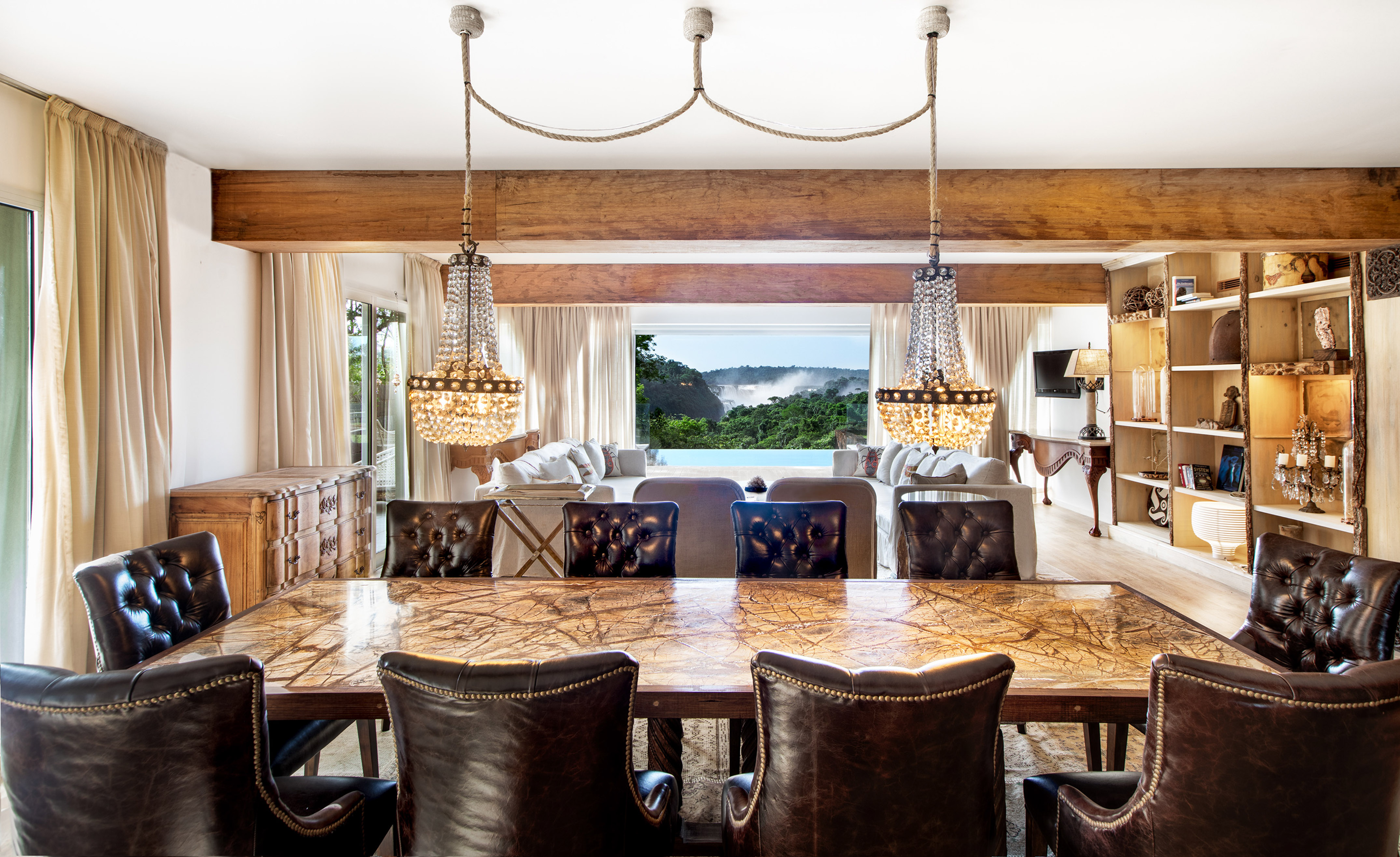 Dining room with marble table, leather chairs, crystal chandeliers, and waterfall view.