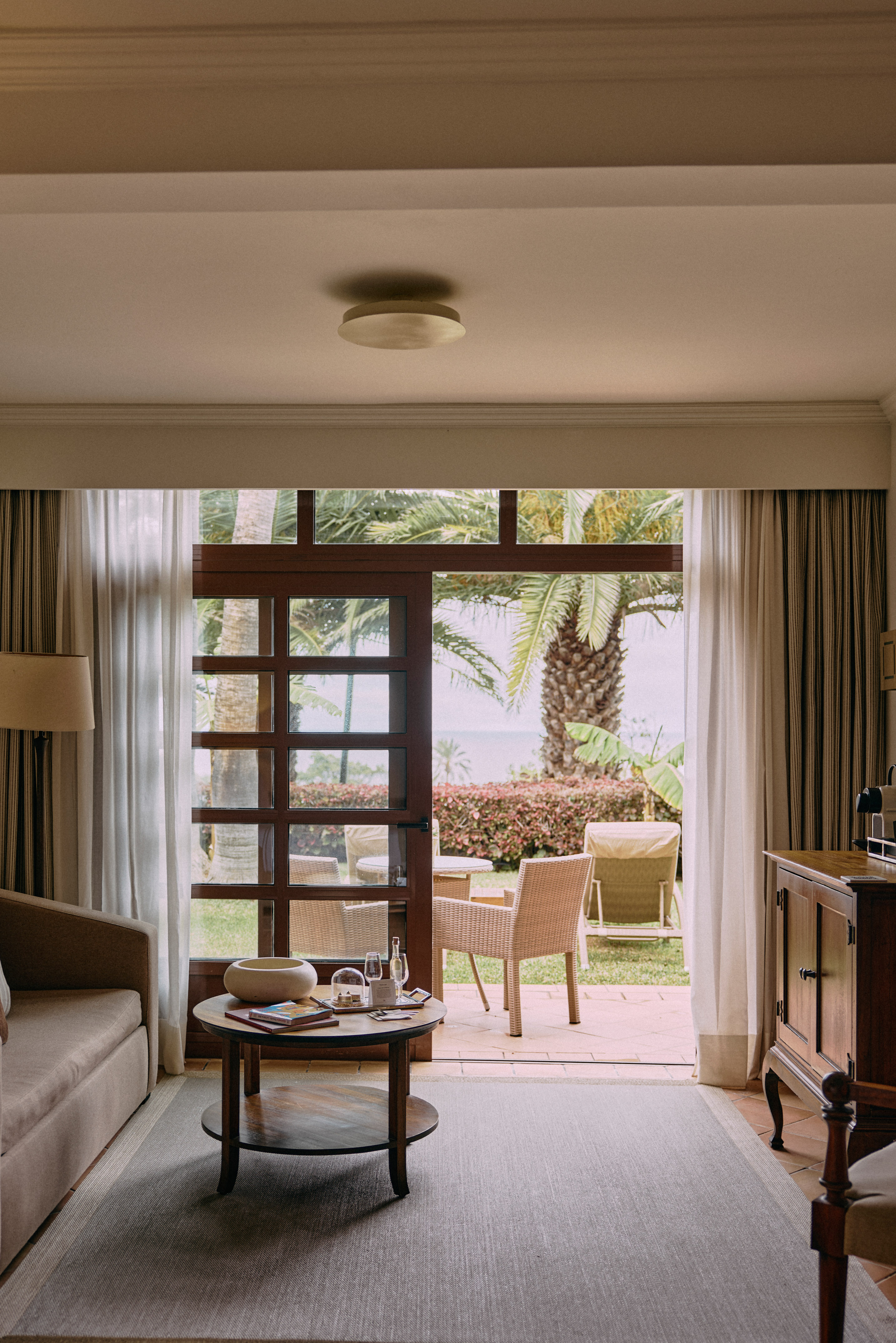 Hotel living room opening to a tropical garden patio with palm trees and sea.