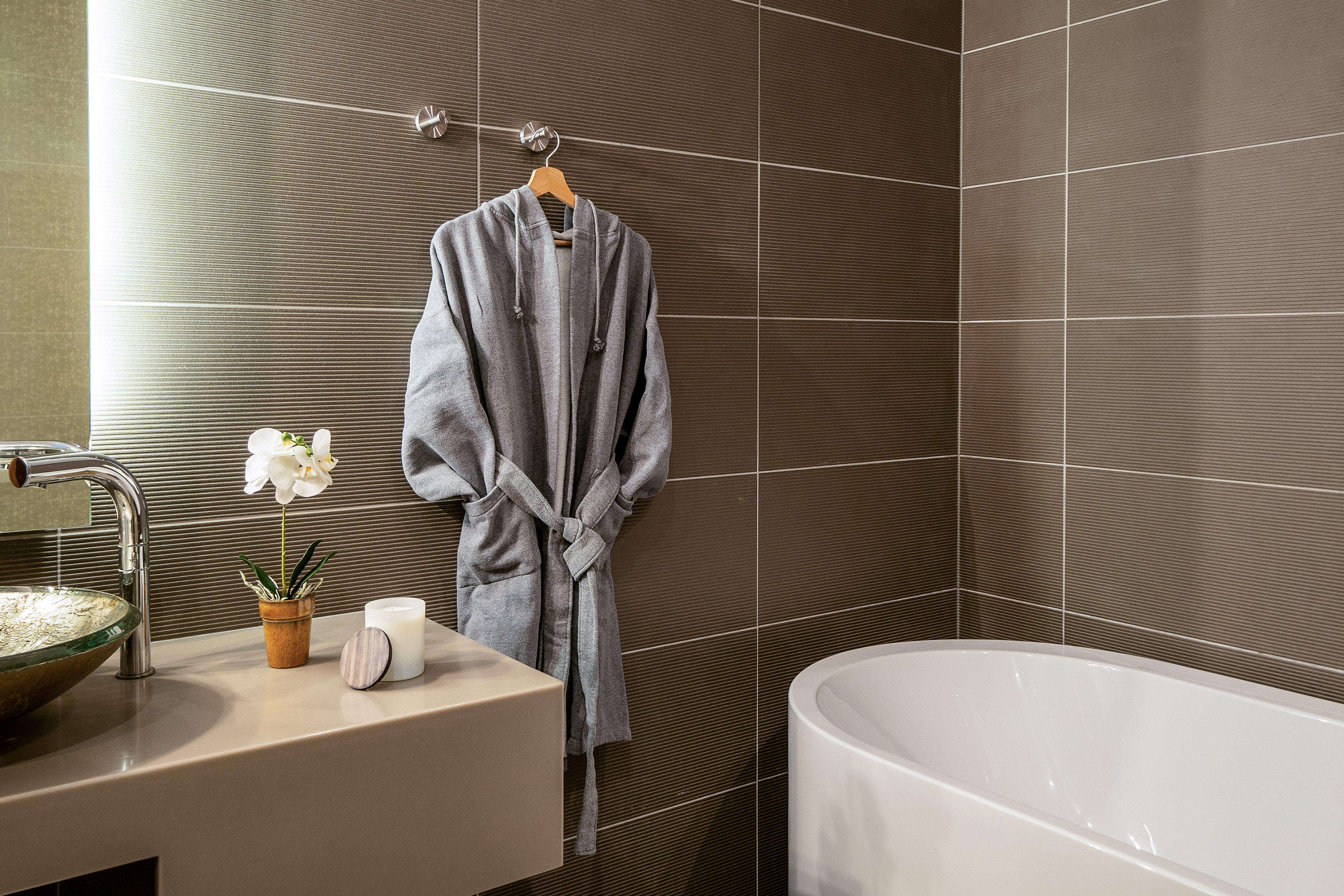 Modern bathroom with gray bathrobe, white bathtub, and brown textured tiles.