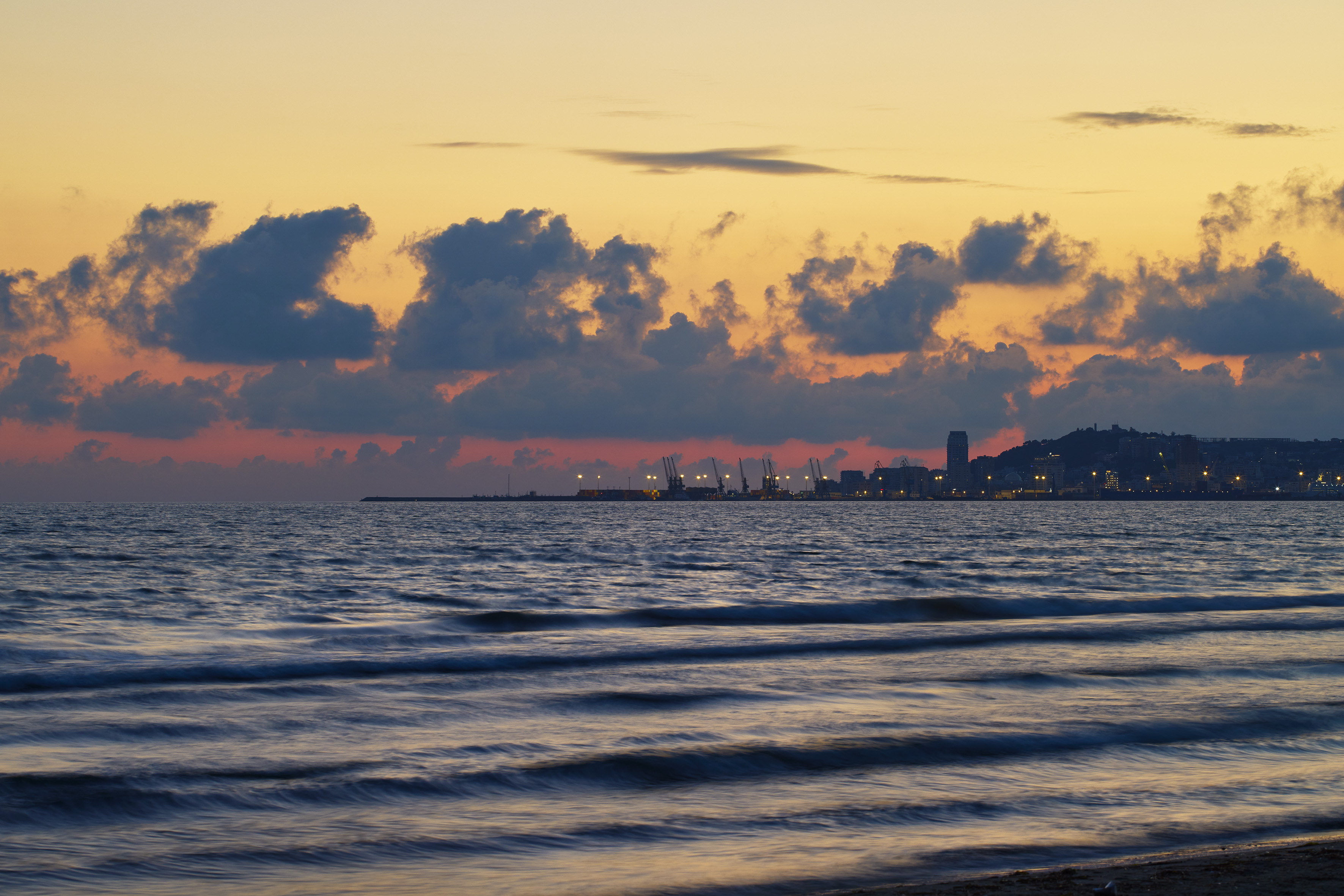 Sunset over wavy ocean, distant city skyline with port cranes.