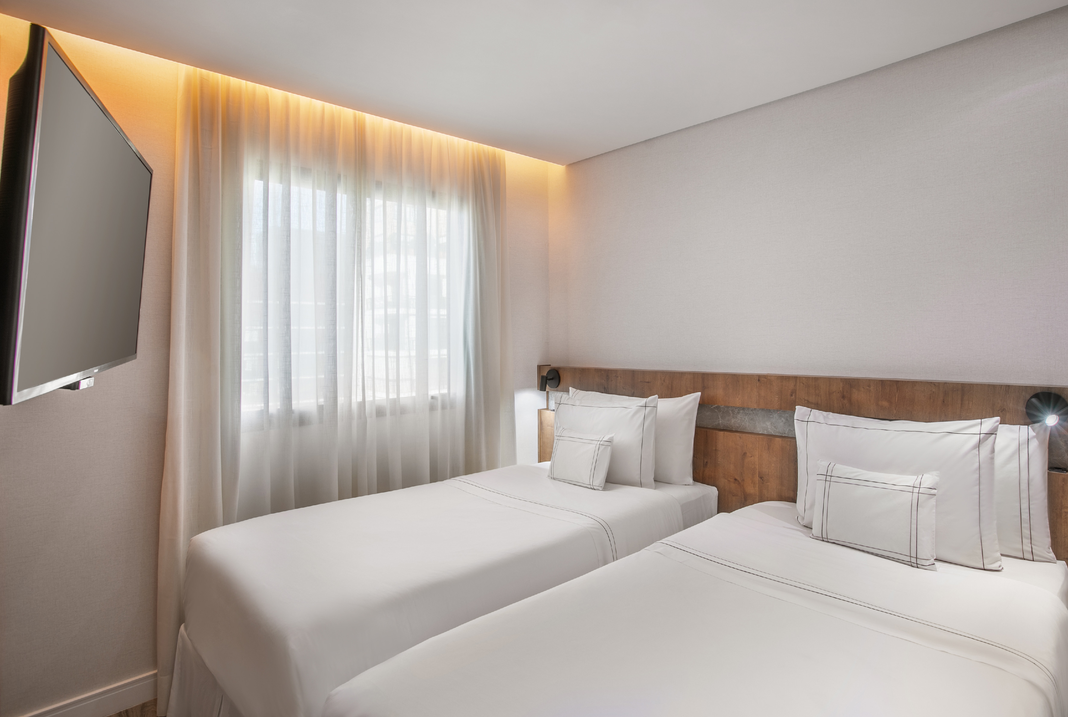 Bright hotel room with two twin beds, wall-mounted TV, and sheer curtains.
