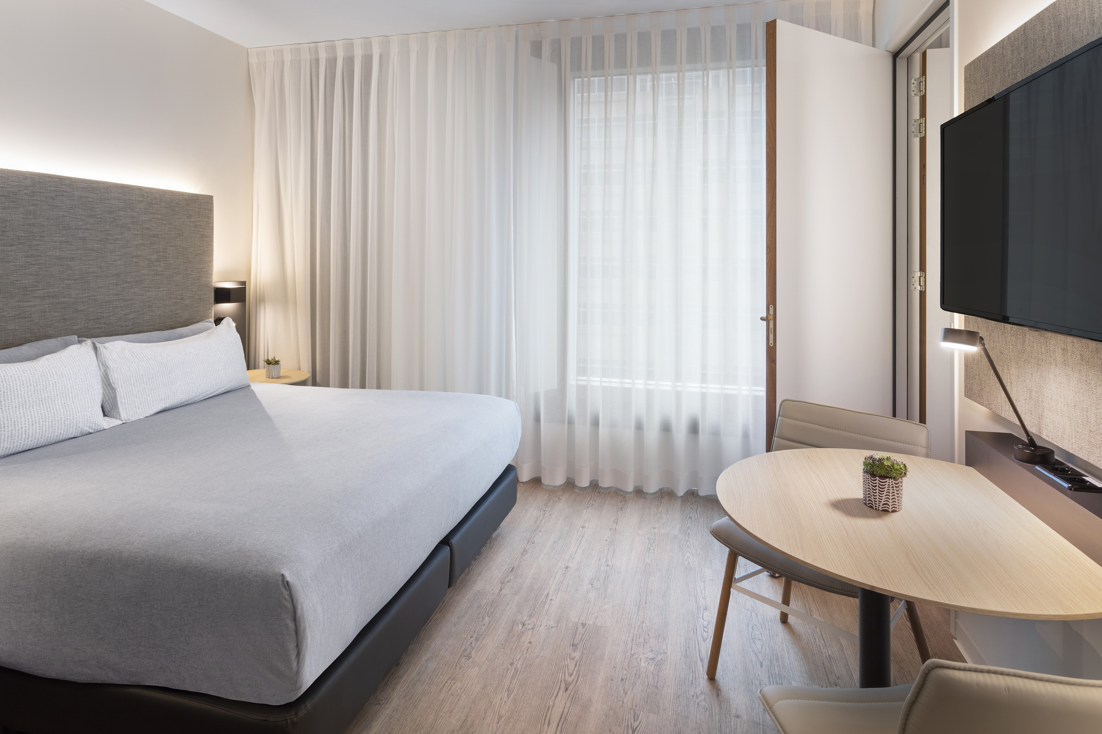 Modern hotel guest room with gray bed, sheer curtains, wood floor, and dining table.