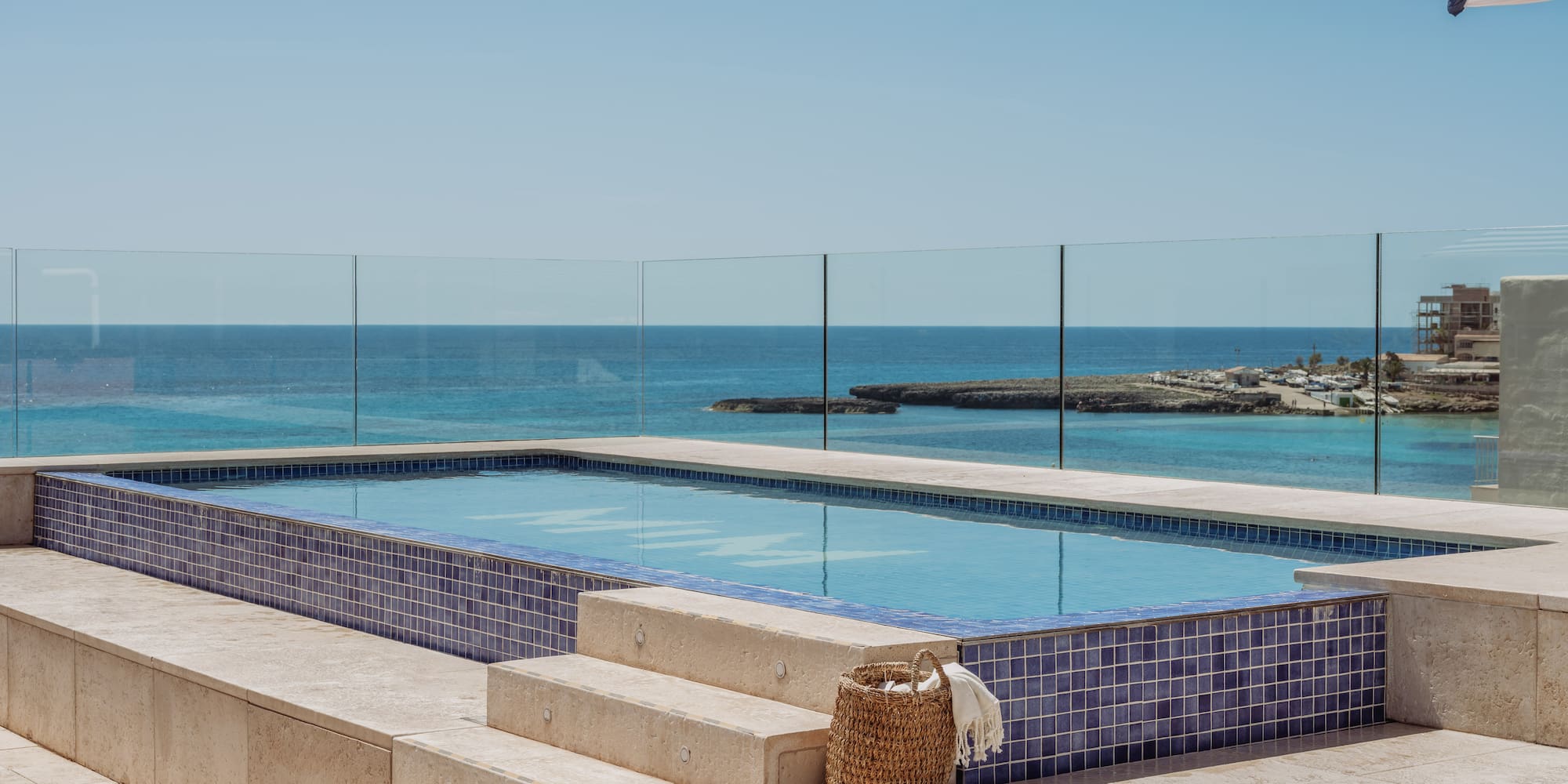 Piscina azul con vista al mar, terraza y sombrilla rayada.