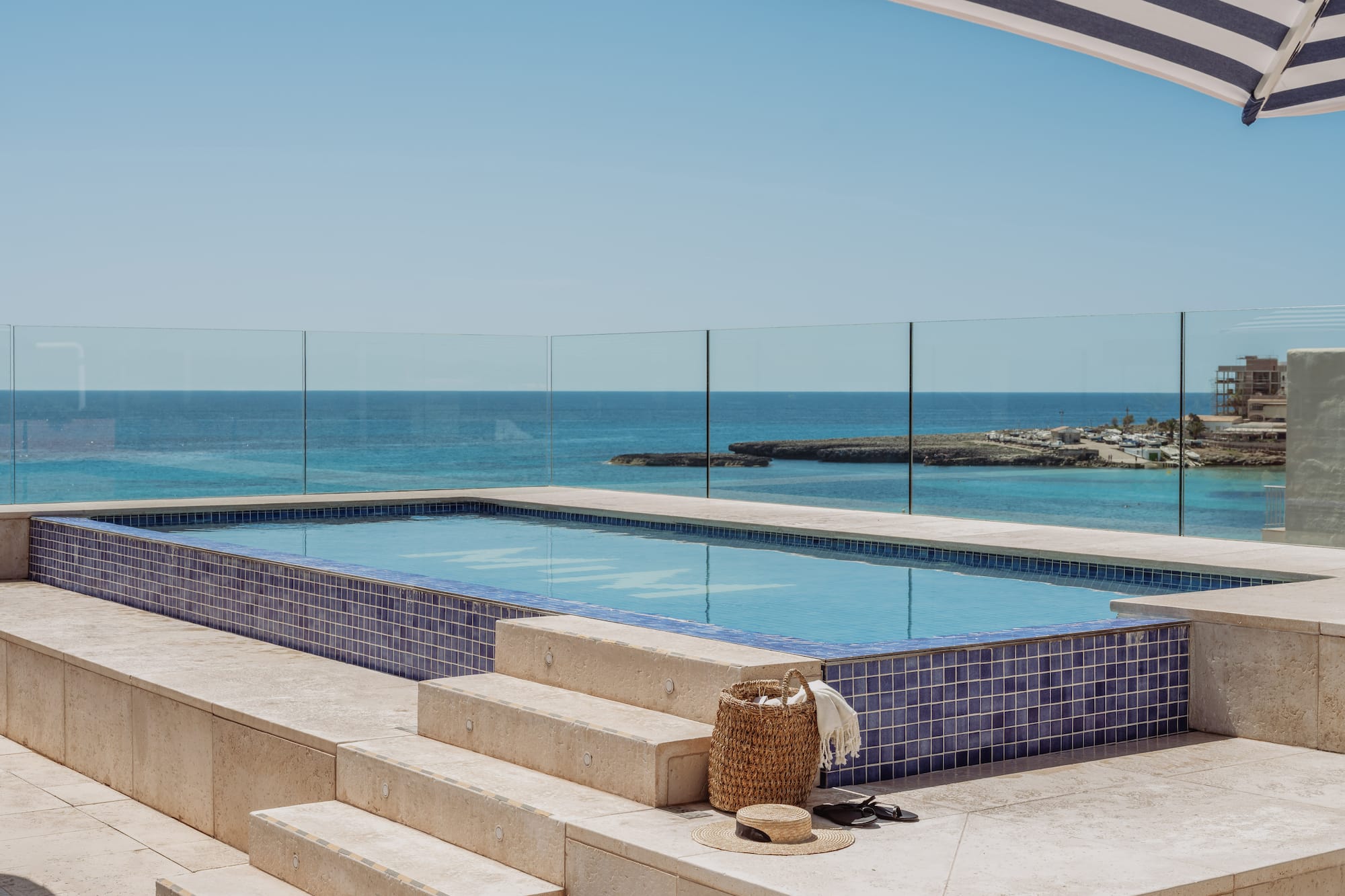 Piscina azul con vista al mar, terraza y sombrilla rayada.