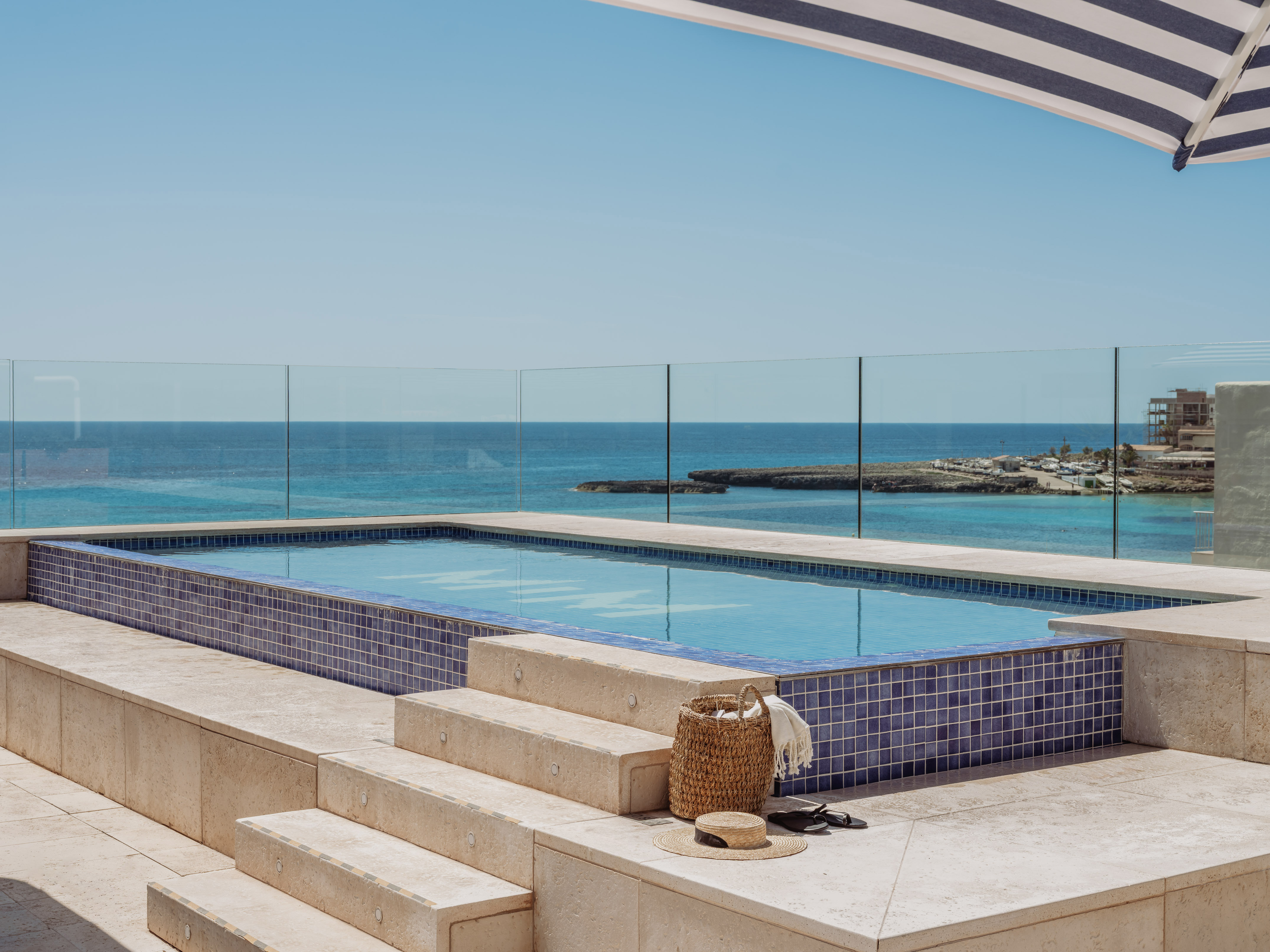 Piscina azul con vista al mar, terraza y sombrilla rayada.