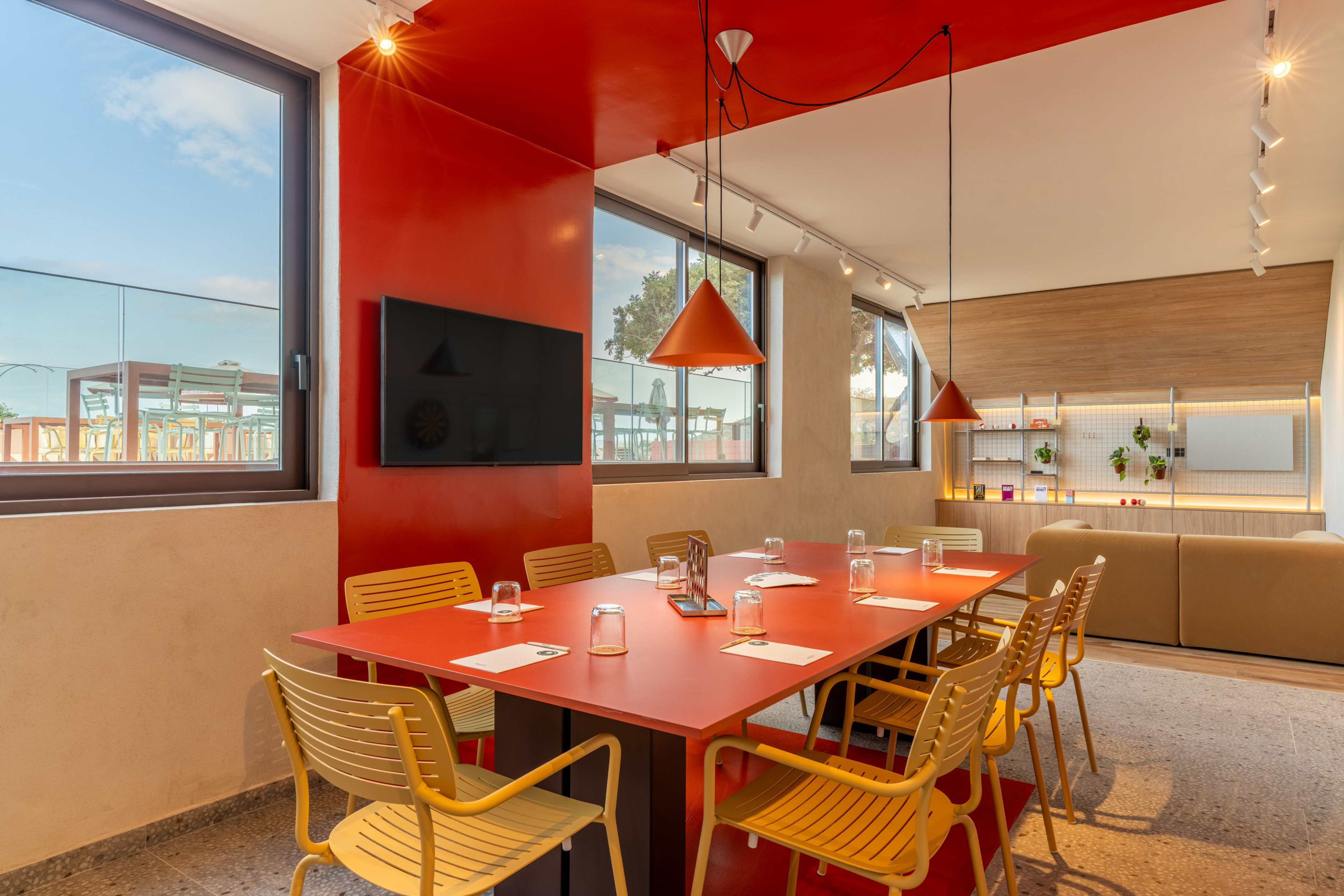 Vibrant meeting room with red table, yellow chairs, TV, and large windows.