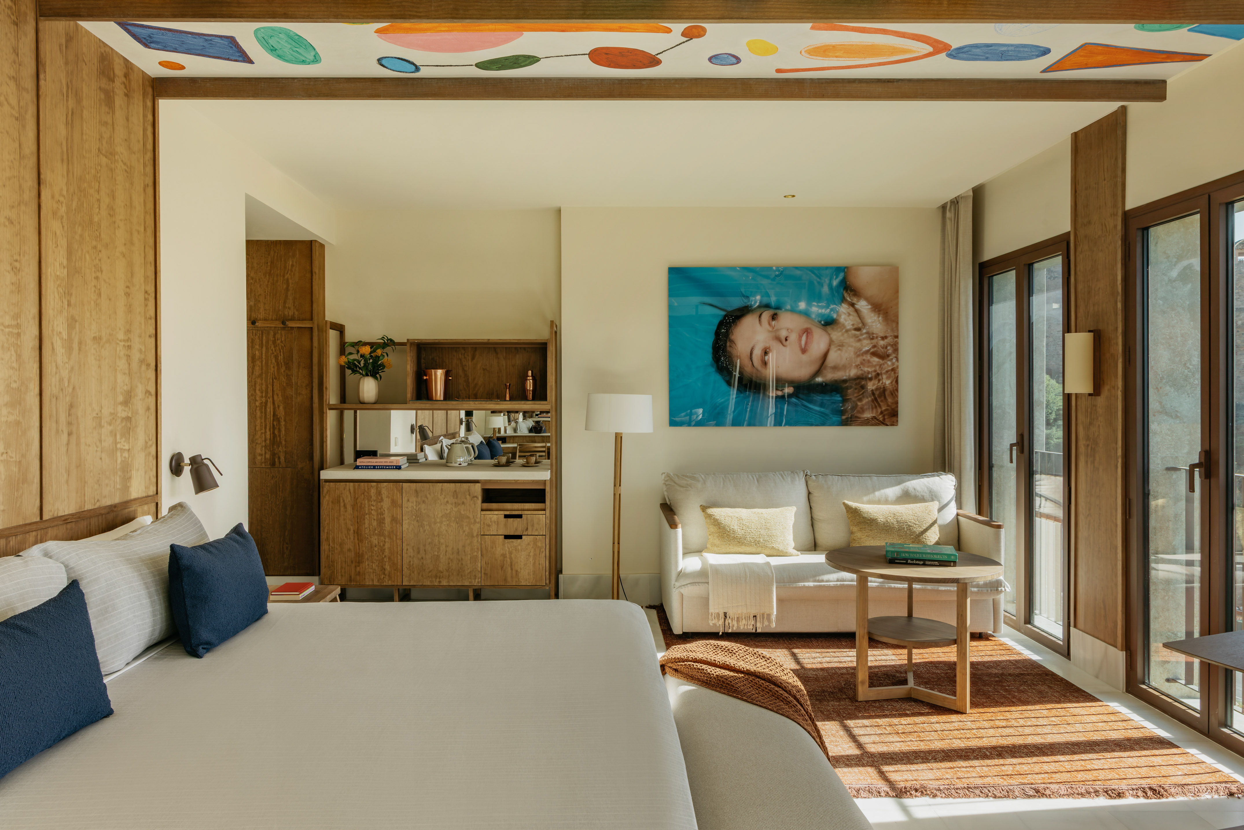Modern hotel room with wood paneling, large bed, sofa, and colorful abstract ceiling.