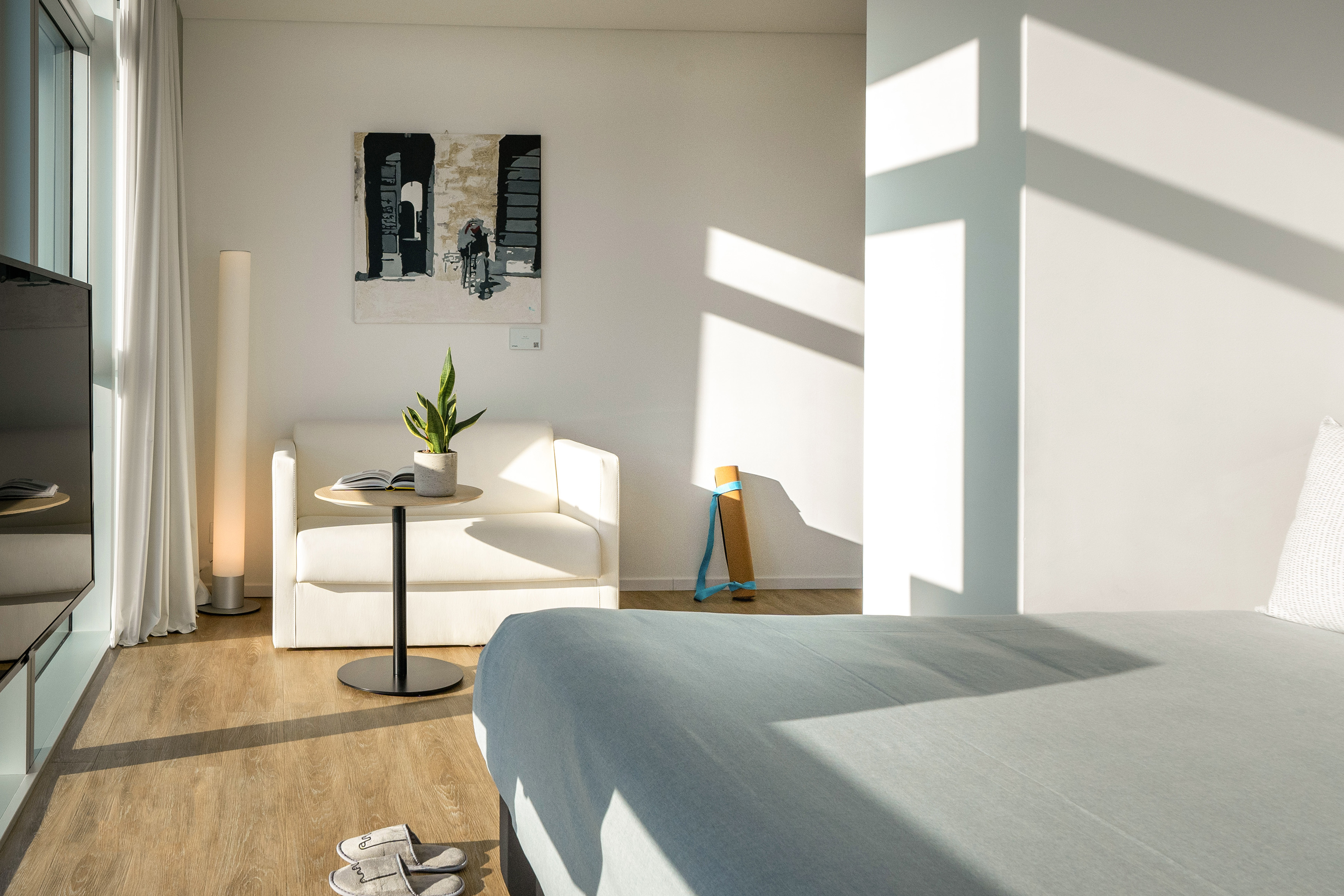 Modern bedroom with white sofa, abstract art, sunlit wood floor, and yoga mat.