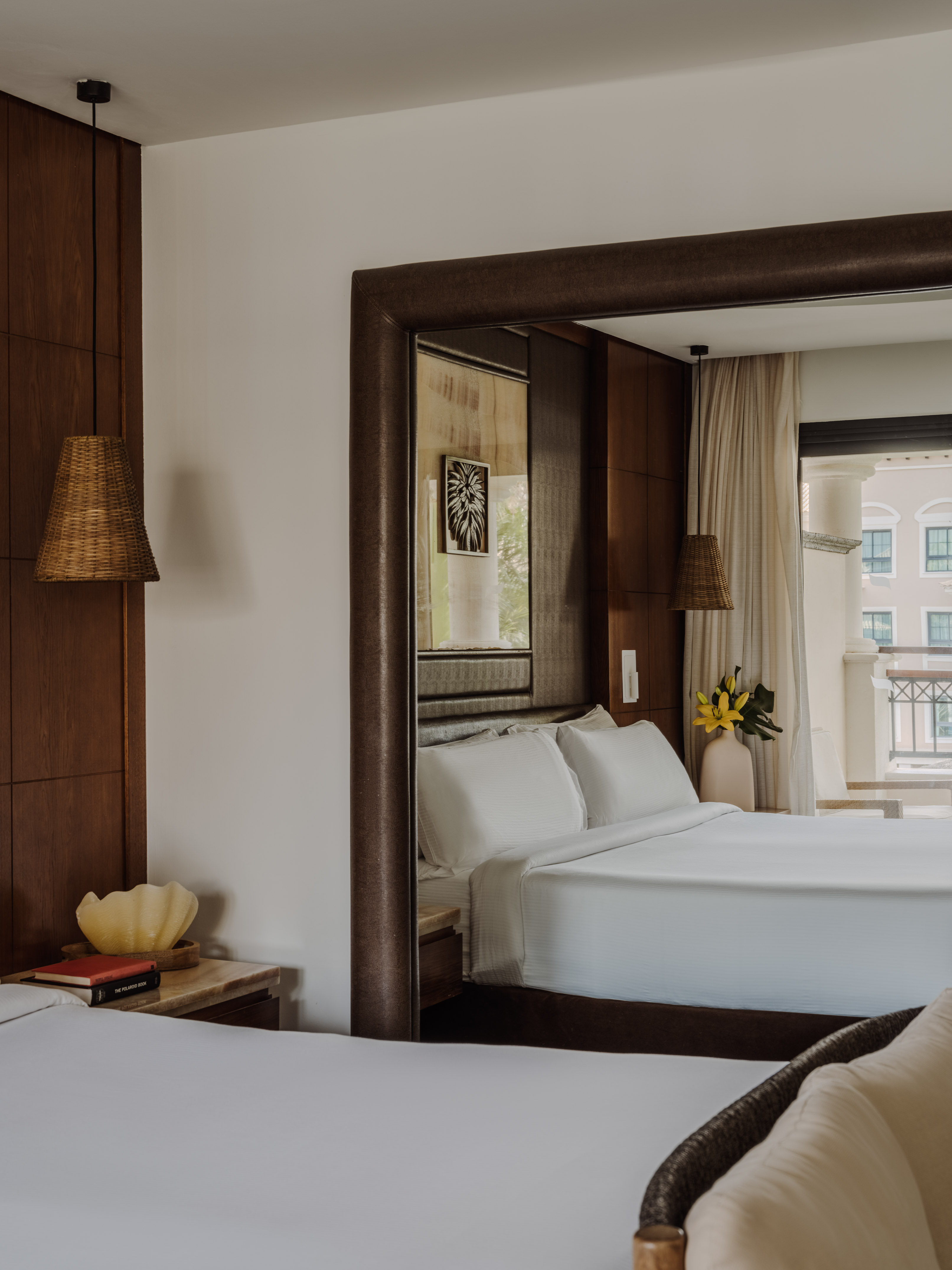 Elegant hotel bedroom with two beds, large mirror, woven lamp, and balcony view.