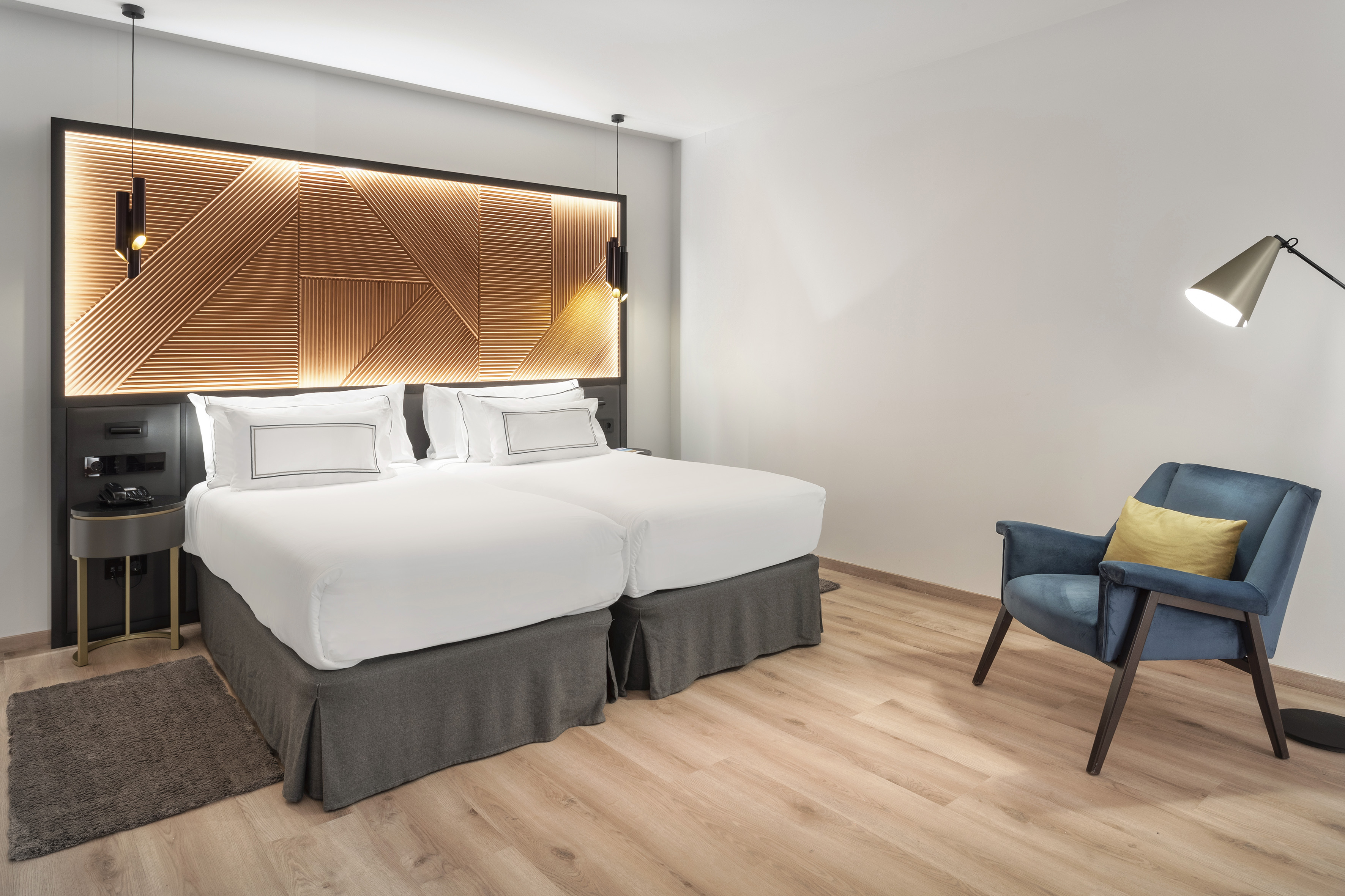 Modern hotel room: twin beds, backlit headboard, blue armchair, and wood floor.